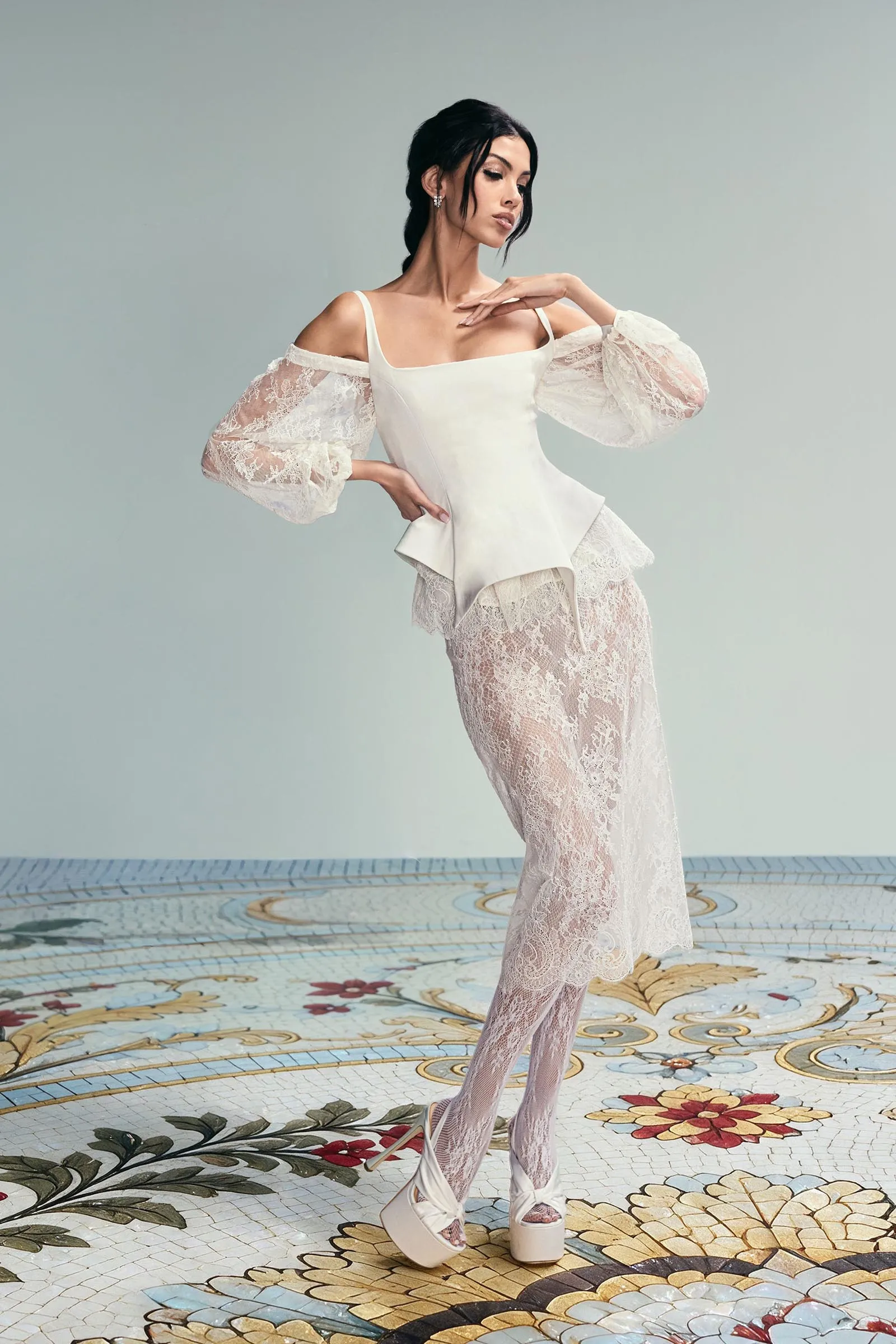 Model in a white lace off-shoulder gown with voluminous sheer sleeves, standing on a colorful floral mosaic floor.