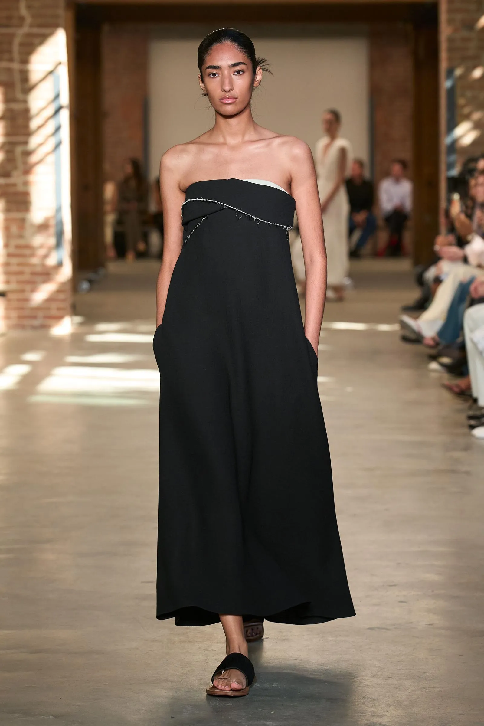 Model walks a runway in a strapless black dress with a curved white underlayer and stitched metallic chain detail across the bodice.