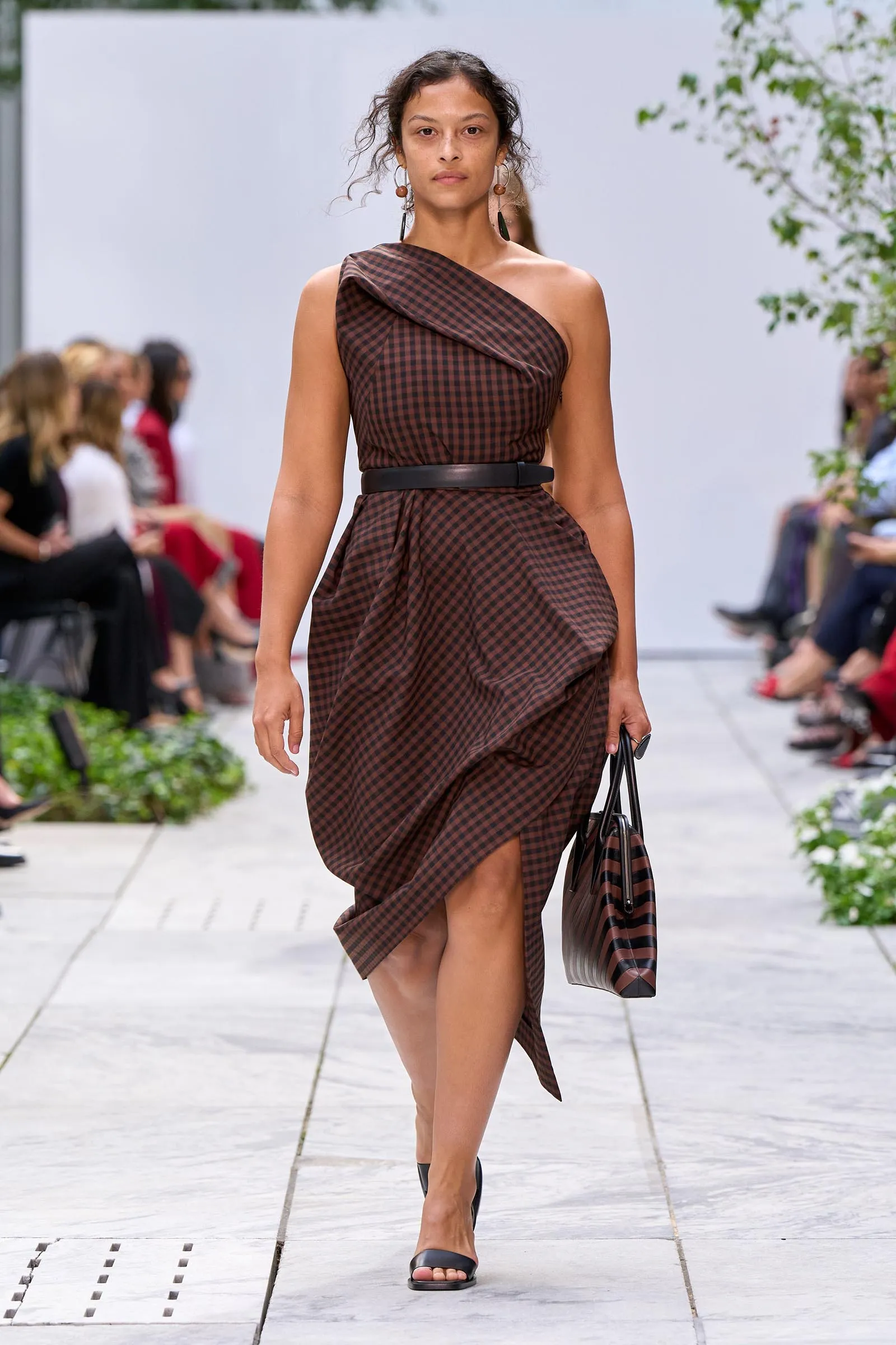 Model walking a fashion runway in a brown checkered, one-shoulder dress with a black belt and matching striped handbag.