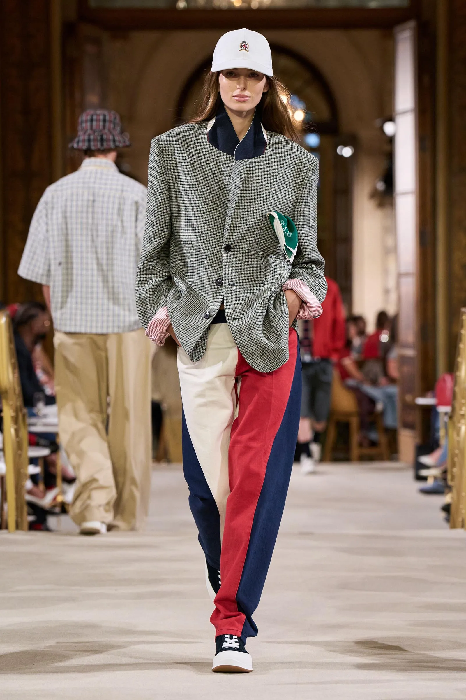 Model walks a runway wearing a gray checkered blazer, navy collar, and color-block red, white, and blue pants, white cap.