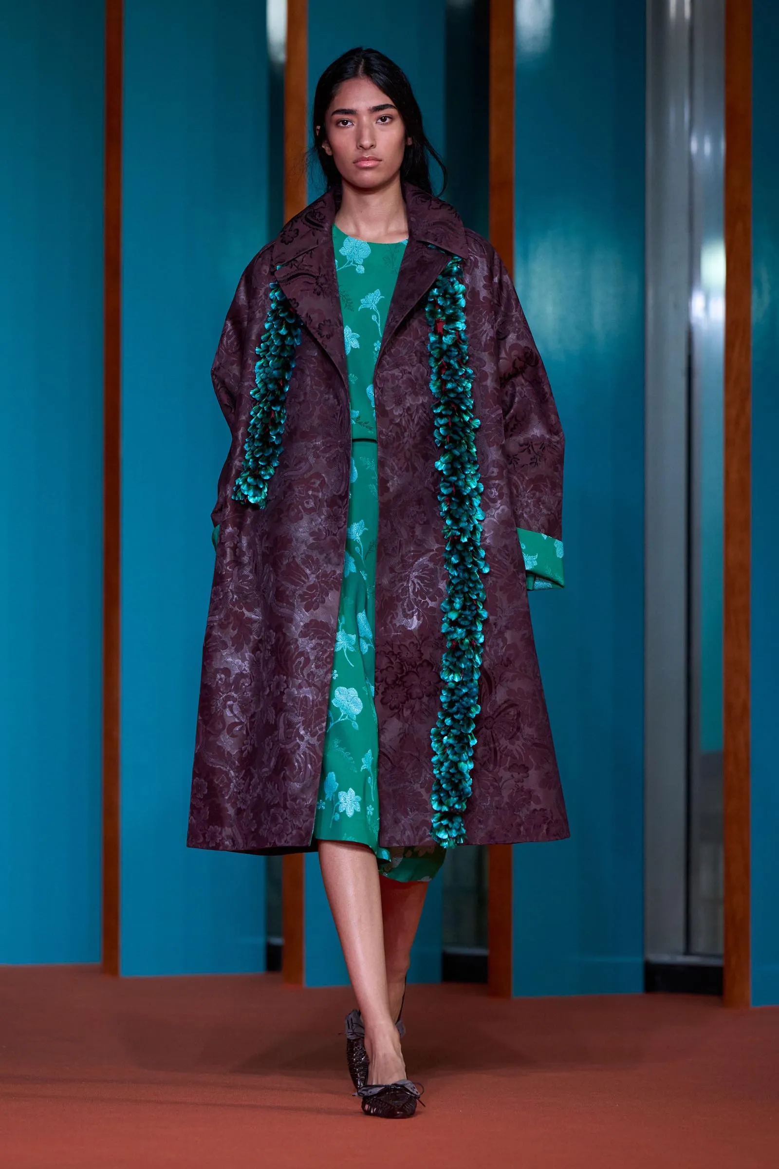 Woman wearing a teal floral dress under a burgundy patterned coat, with teal garland draped over her shoulders, standing in a teal hallway.