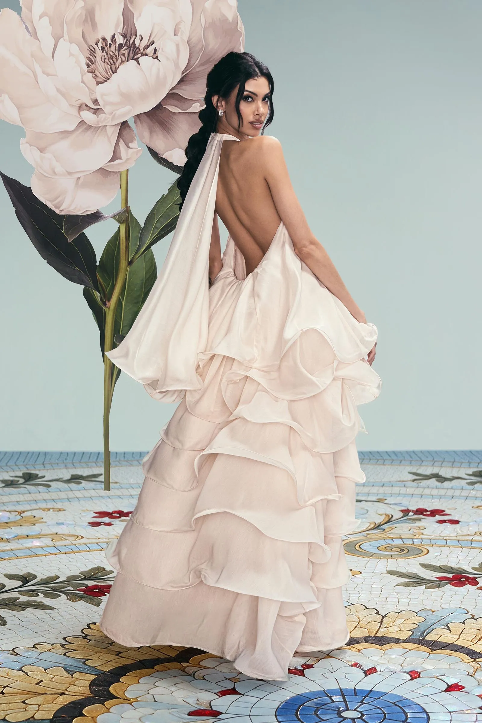 Woman in a backless pale pink gown with a long braid, standing against a large floral backdrop indoors.