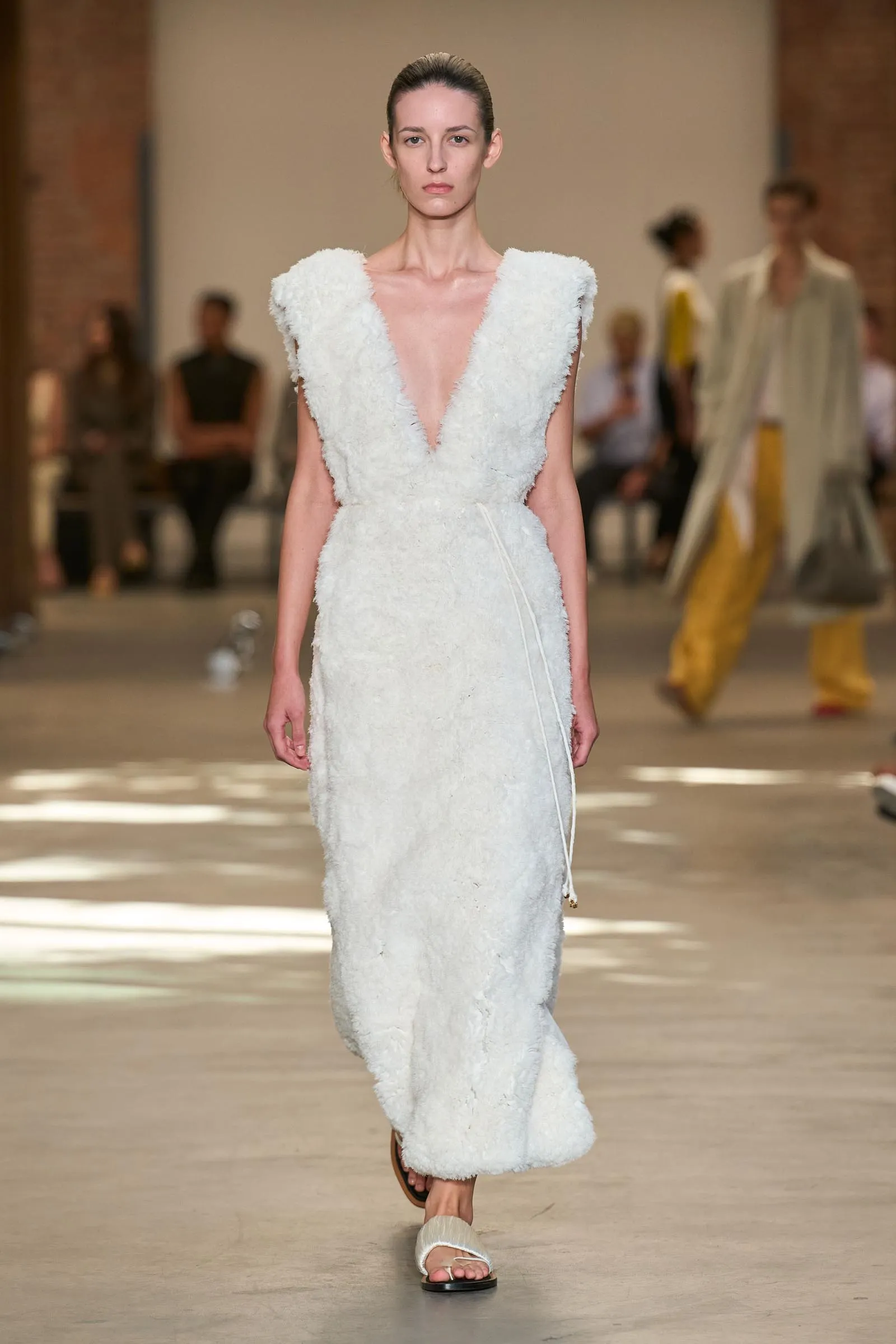 Model walks the runway in a white textured gown with a deep V-neck and wide shoulders, audience blurred in the background on a fashion show.