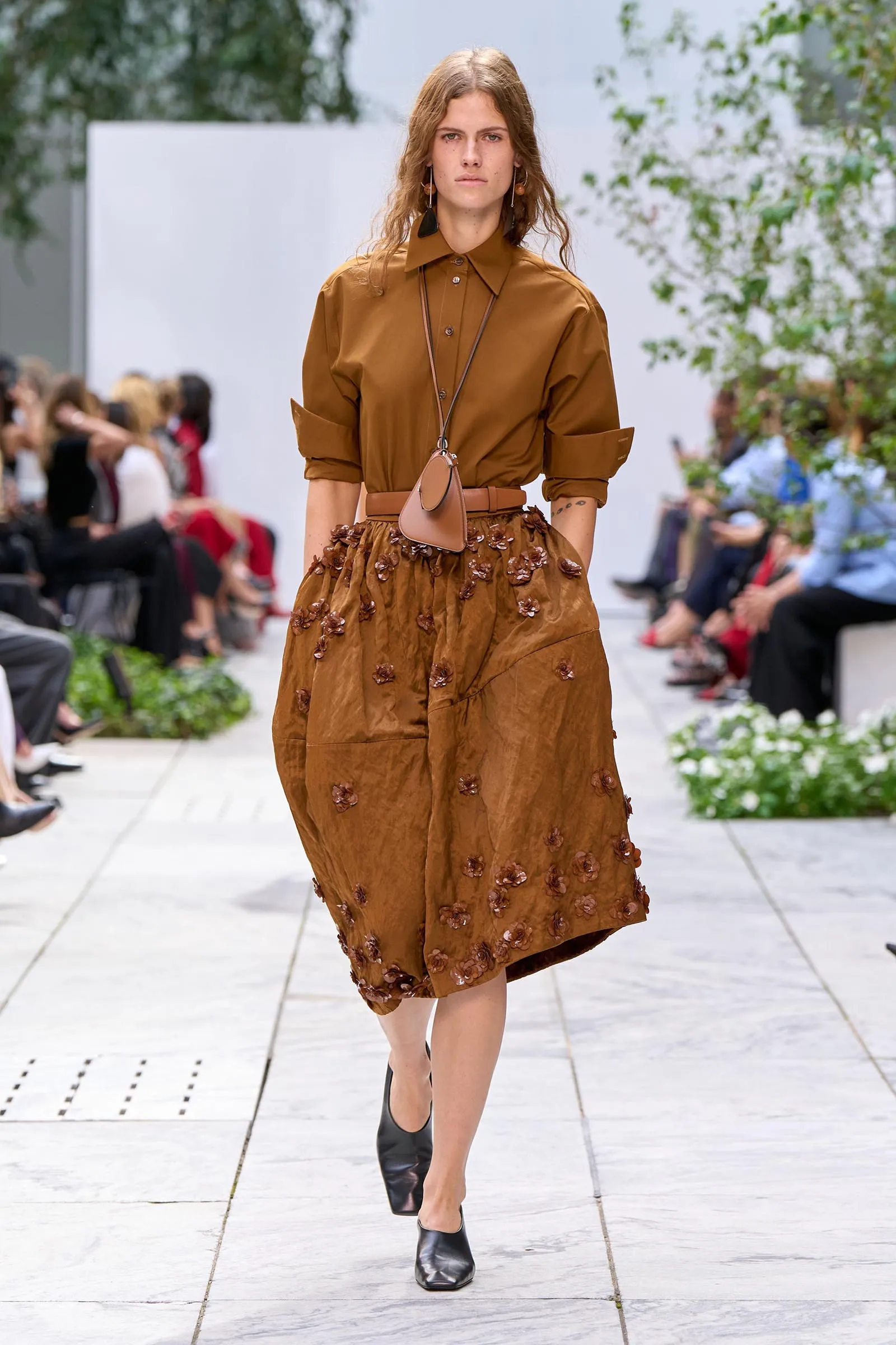 Model walking a fashion show wearing a brown button-up blouse and a brown flower-embellished skirt, with a small tan bag hanging from a long strap.