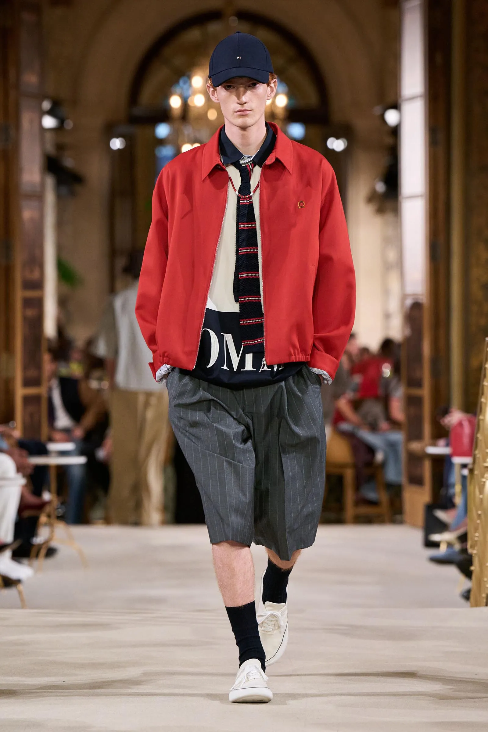 Male fashion model walking a runway in a red jacket, striped tie, dark shorts, and navy cap; audience blurred in the background.