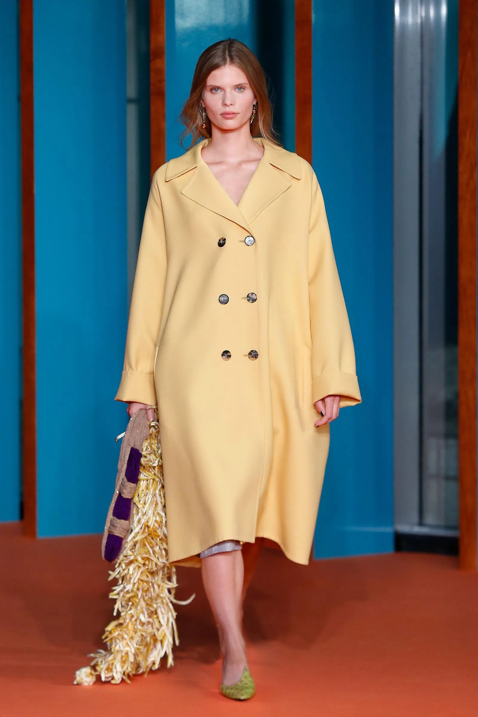 Model walking a fashion runway in a pale yellow double-breasted coat, holding a patterned fringed scarf, with green shoes against blue panels.