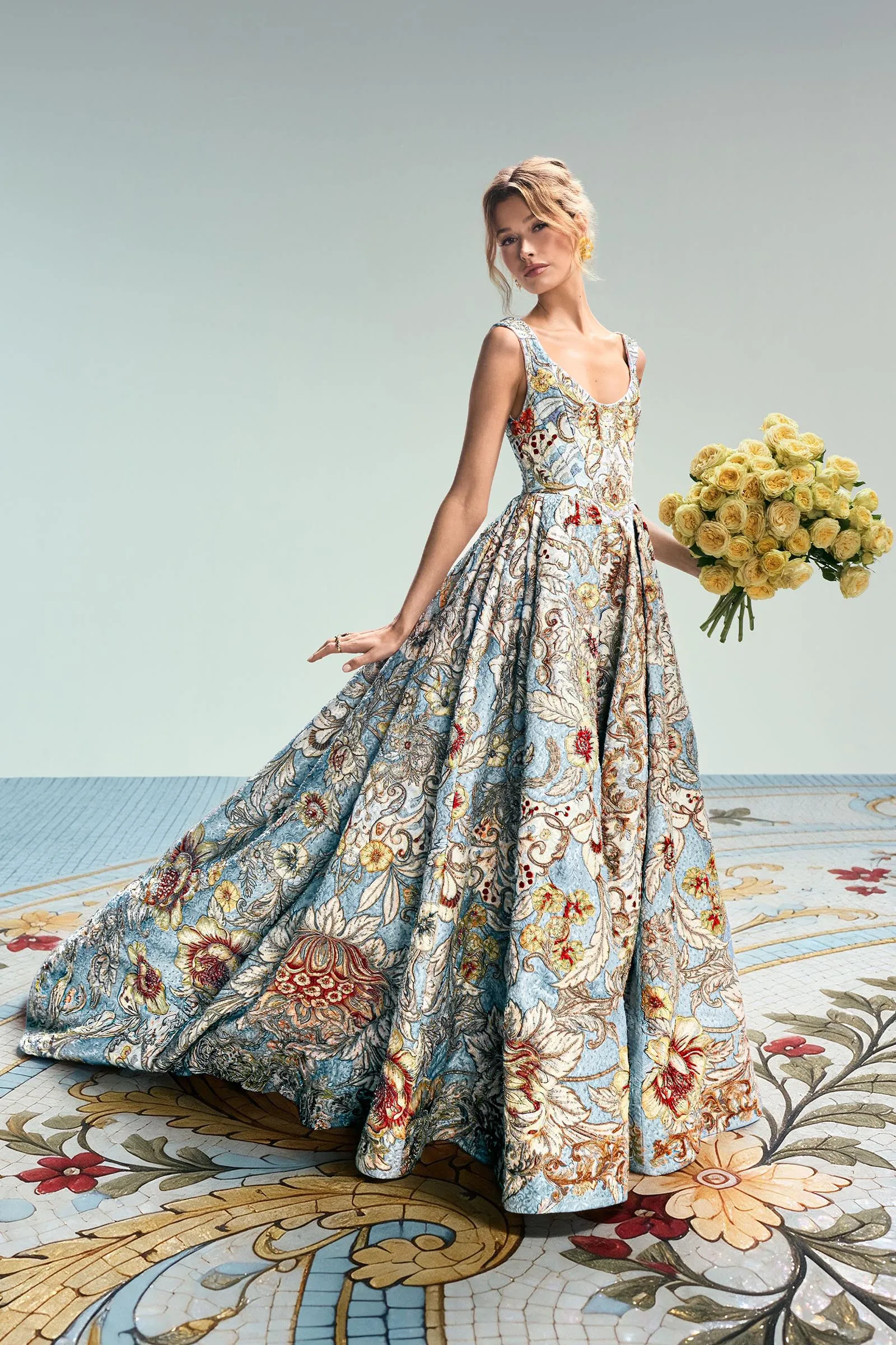 Model wearing a detailed blue and gold embroidered gown, holding a large bouquet of pale yellow roses.