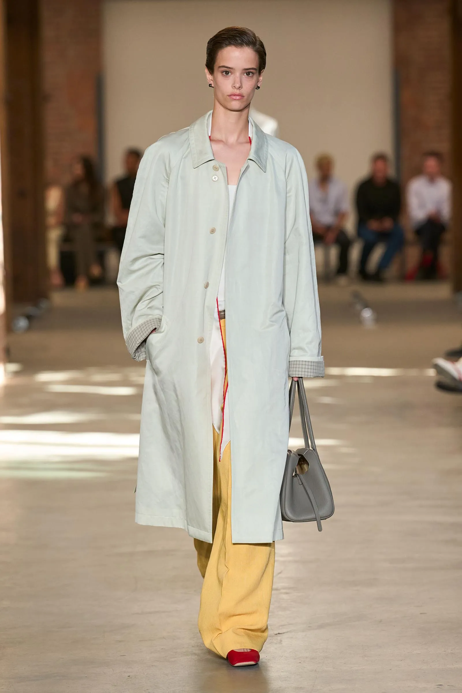 Model walking a runway in a pale gray trench coat, mustard pants, red shoes, and a gray handbag; audience visible in the background.