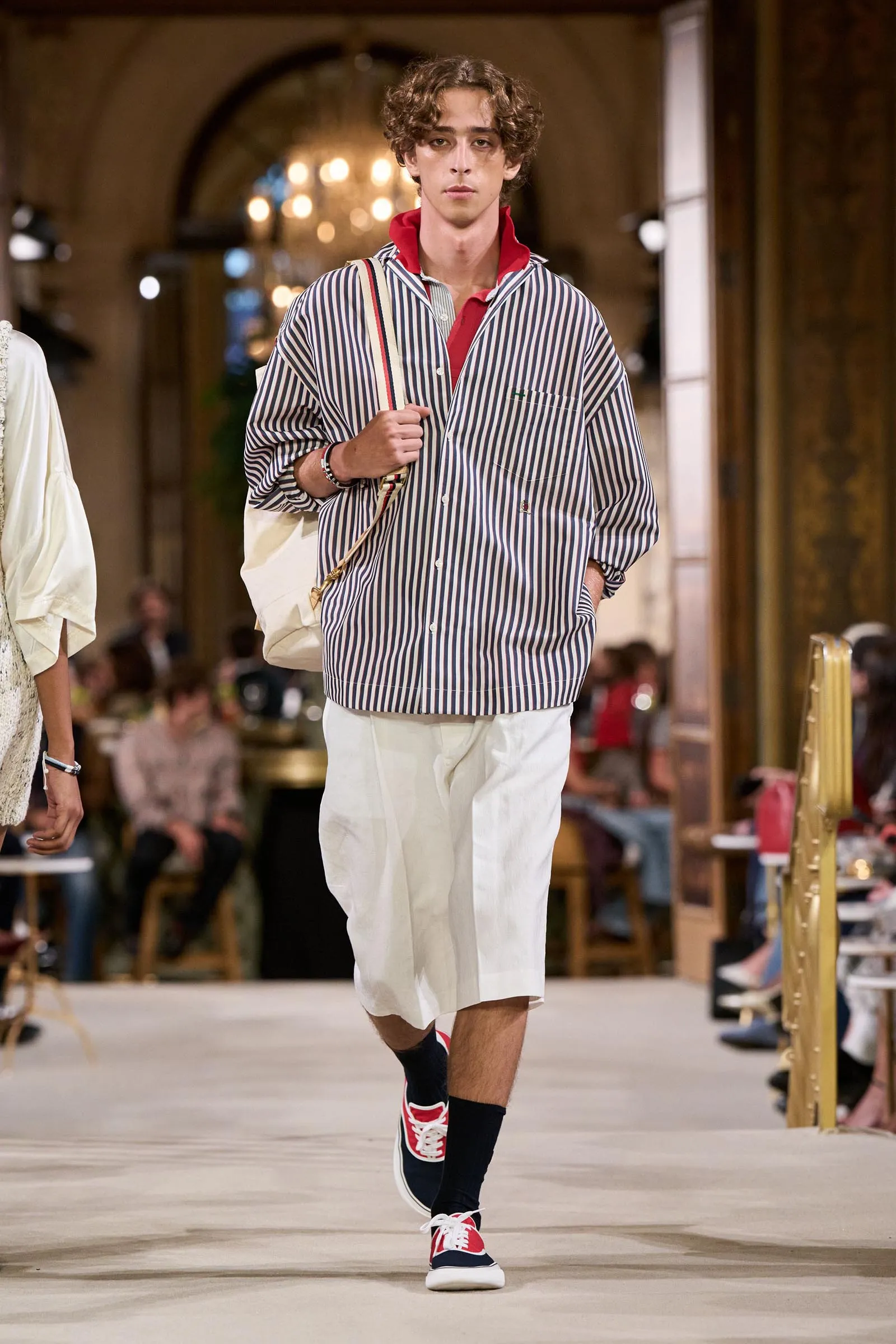 Male model walks a runway in a loose navy-and-white striped shirt with a red collar, white knee-length shorts, and red‑white sneakers.