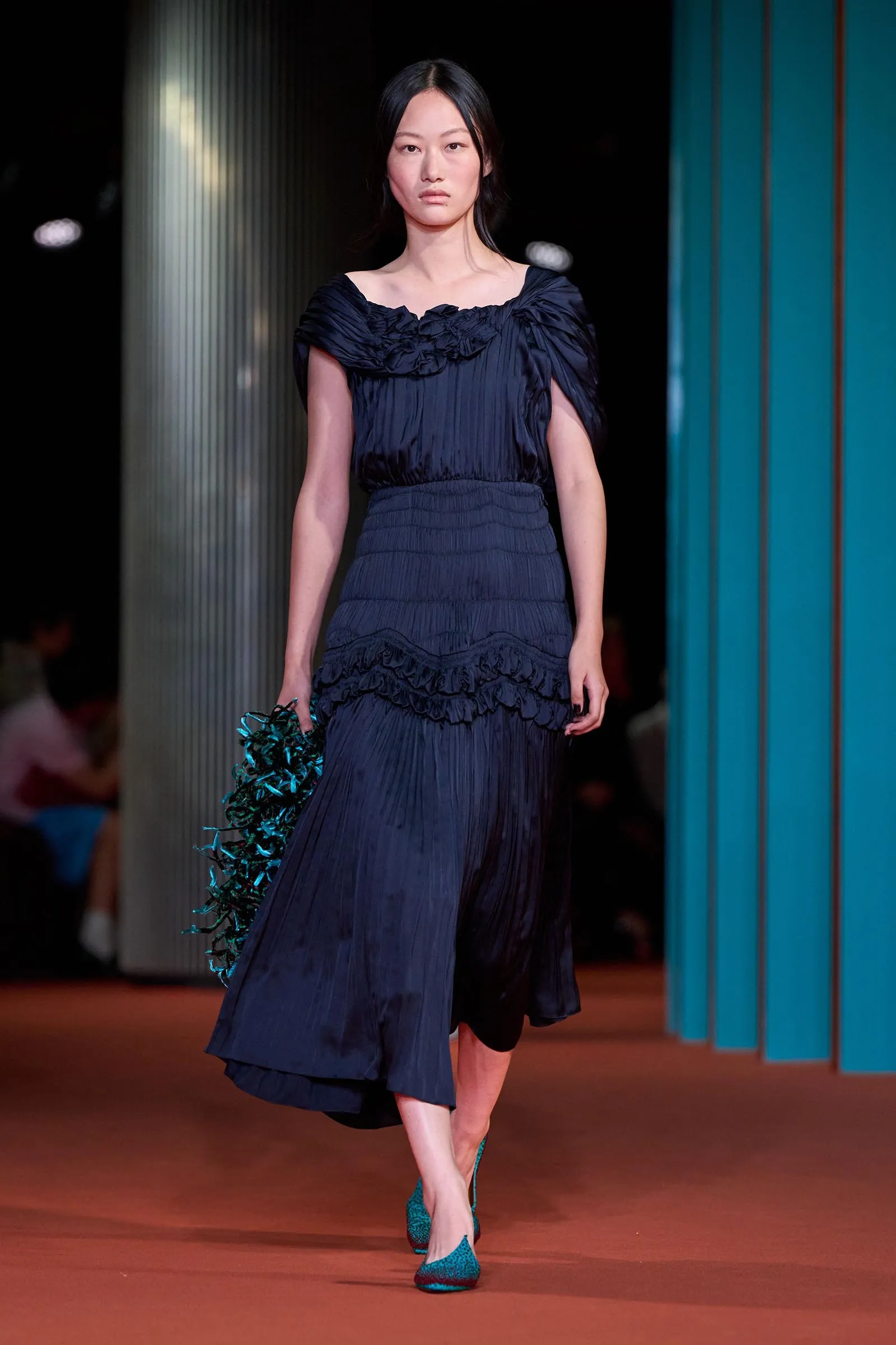 Model walks the runway in a navy blue, pleated dress with ruffled neckline, paired with teal high heels and a teal accessory in hand.