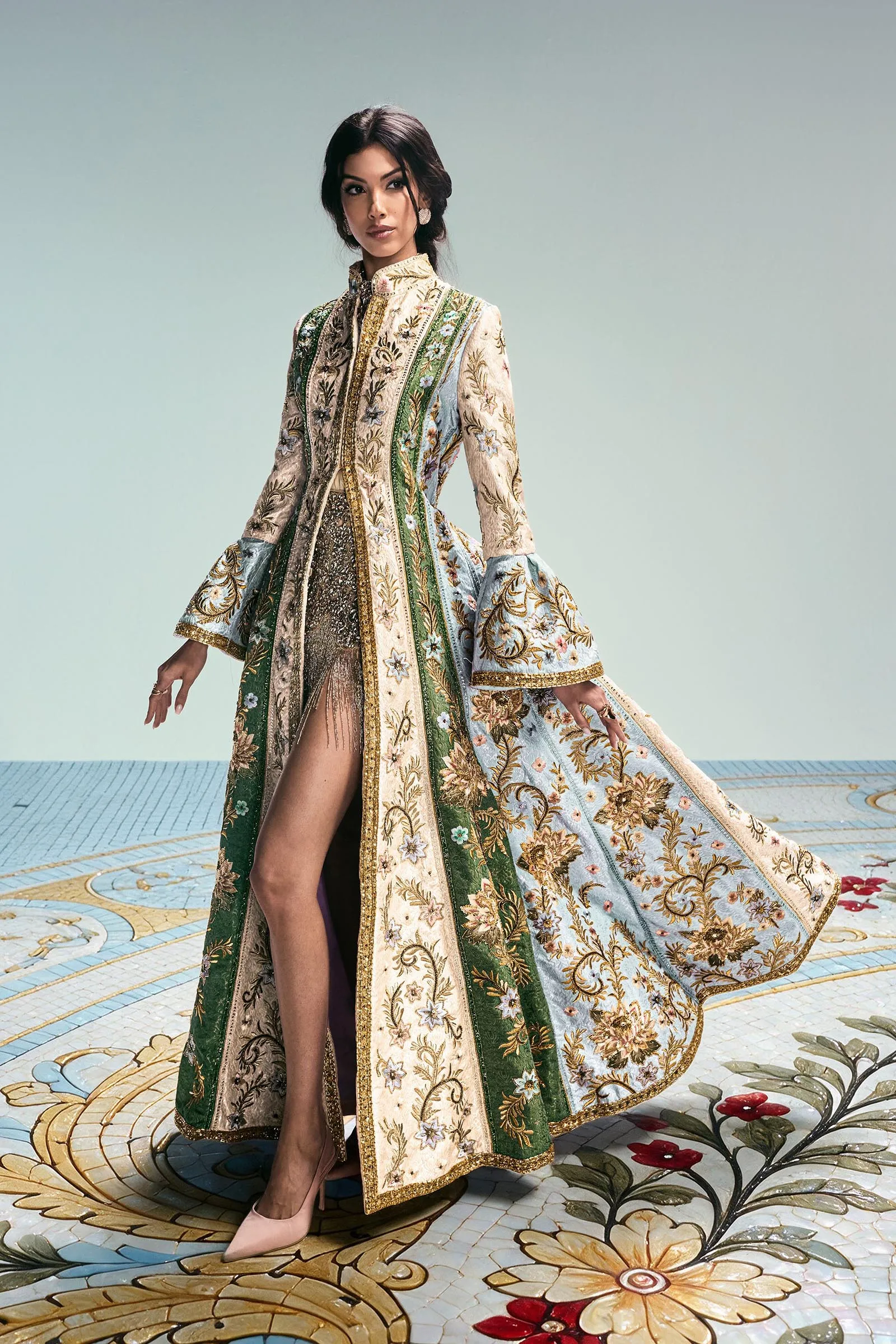 Woman in a richly embroidered cream and green floor-length coat with gold trim, standing on a floral mosaic floor.