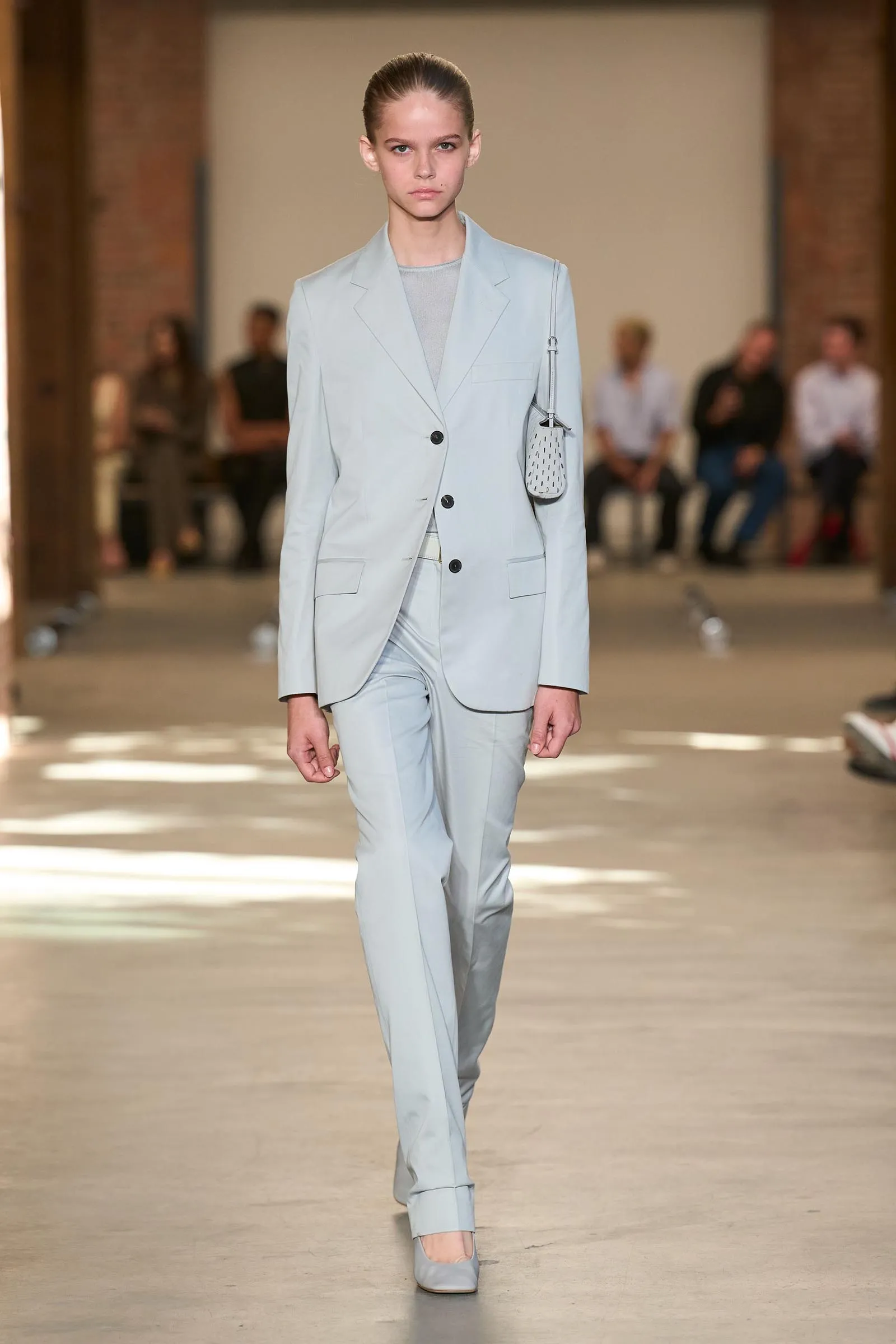 Model walks a fashion runway in a pale blue-gray suit with a matching blazer and trousers, carrying a small white dotted handbag.