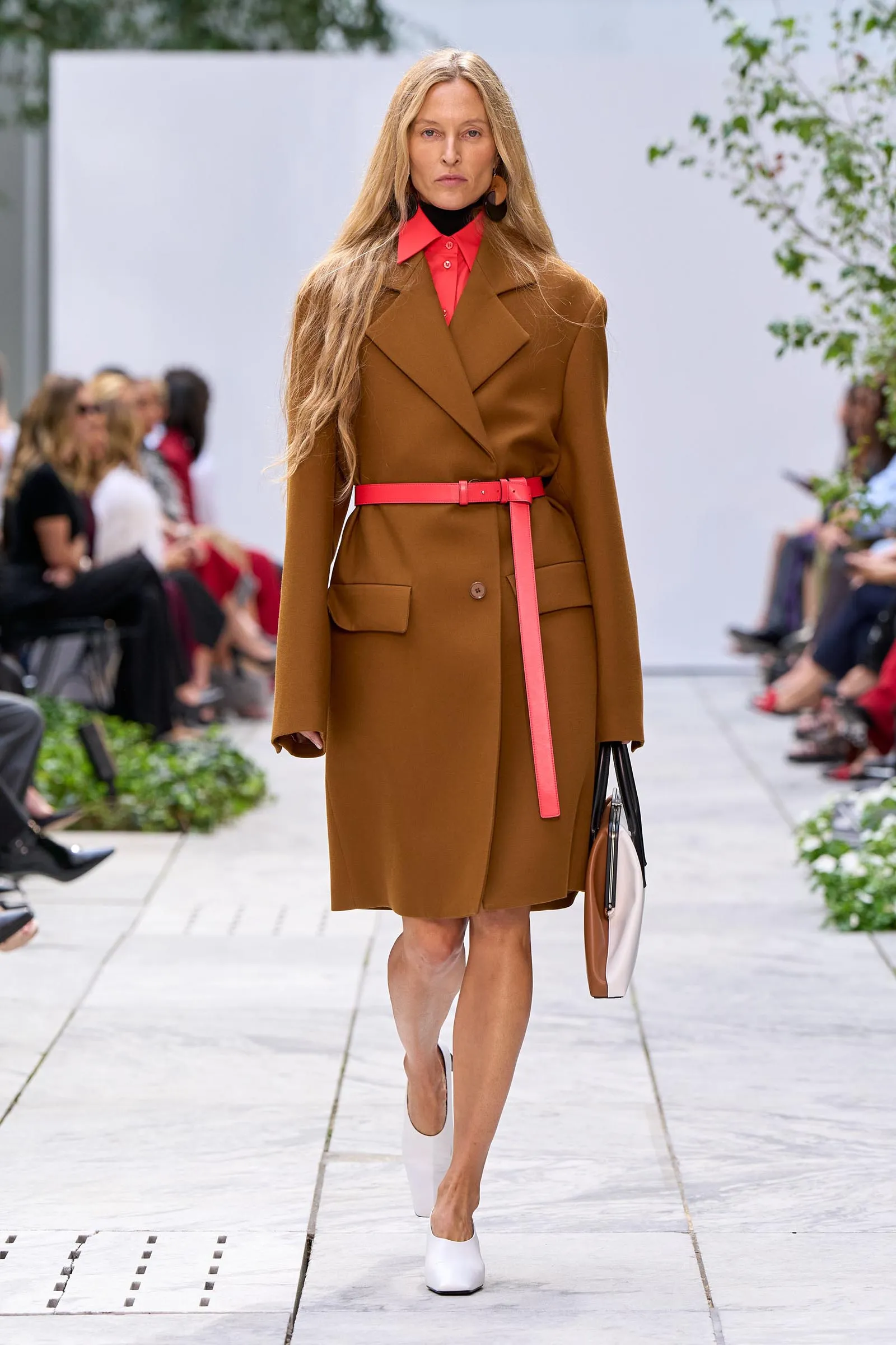Model on an outdoor runway wearing a tan coat with a bright pink belt, red blouse, and white heels while carrying a handbag.