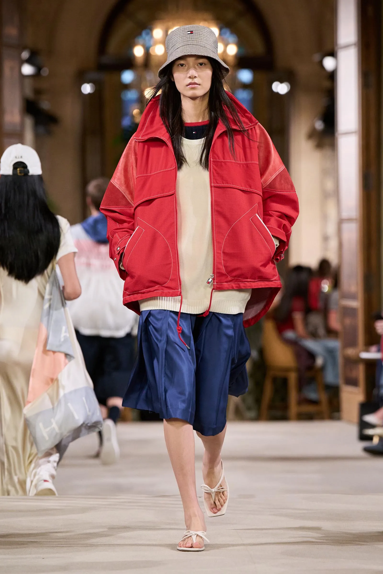Model walks a fashion show wearing a bright red oversized jacket over a beige sweater and blue shorts, plus a checkered bucket hat and white sandals.