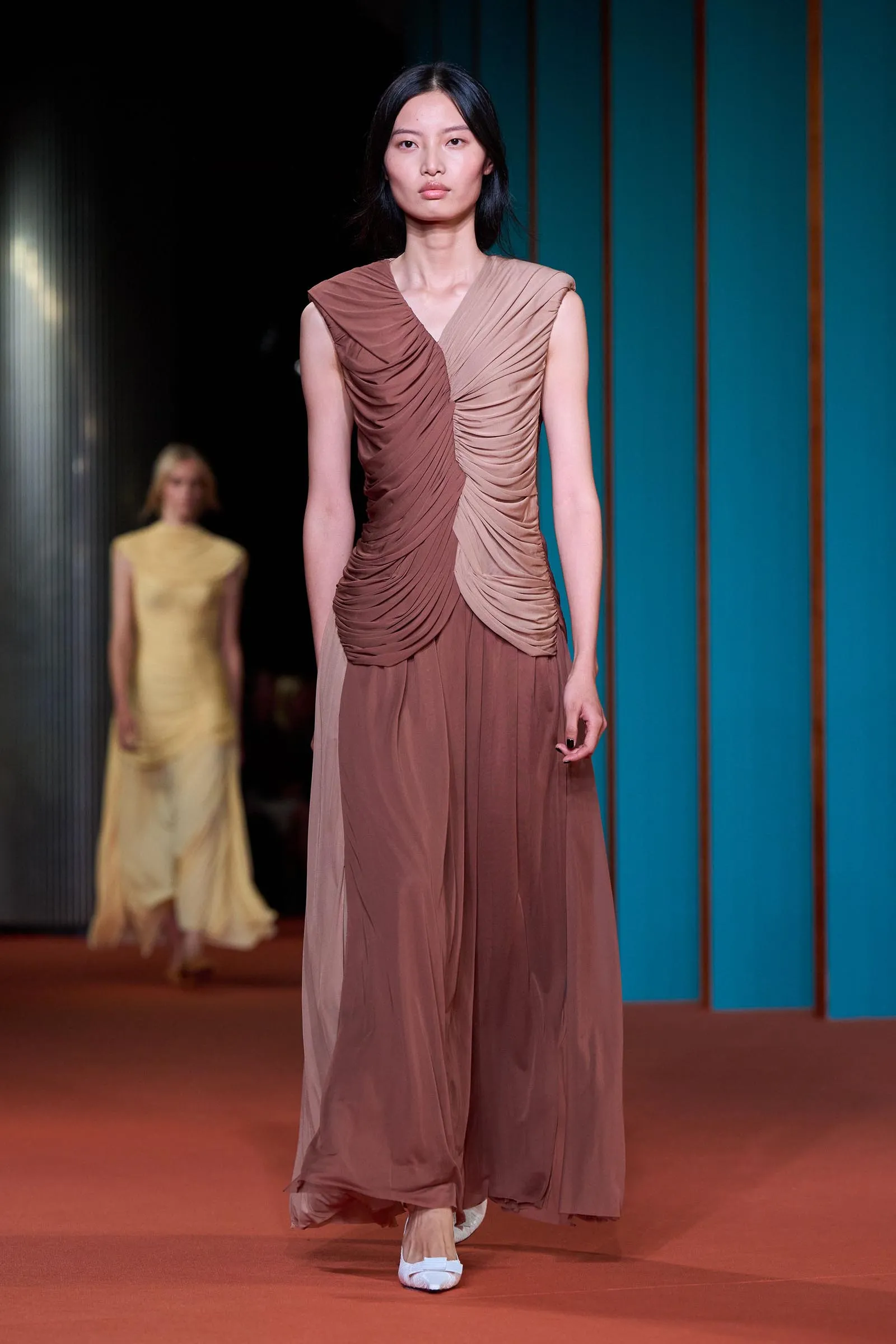 Model walking the runway in a brown ruched, pleated evening gown with a teal backdrop behind her.