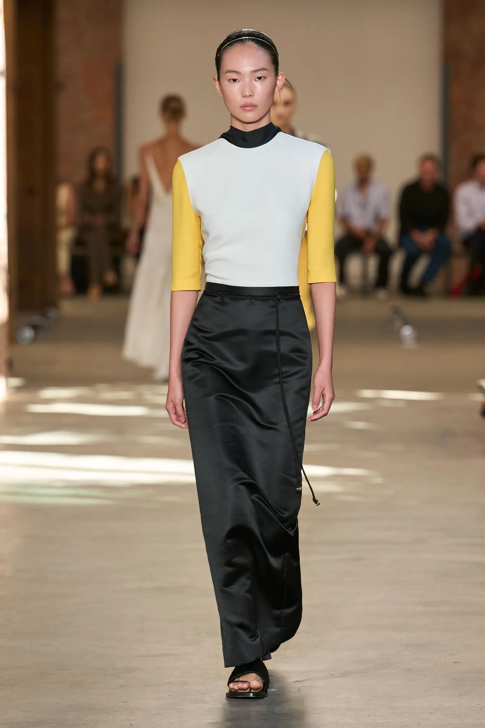 Model walking a fashion runway in a pale blue top with yellow sleeves, black high-waisted satin skirt, and a black underlayer, headband in place.