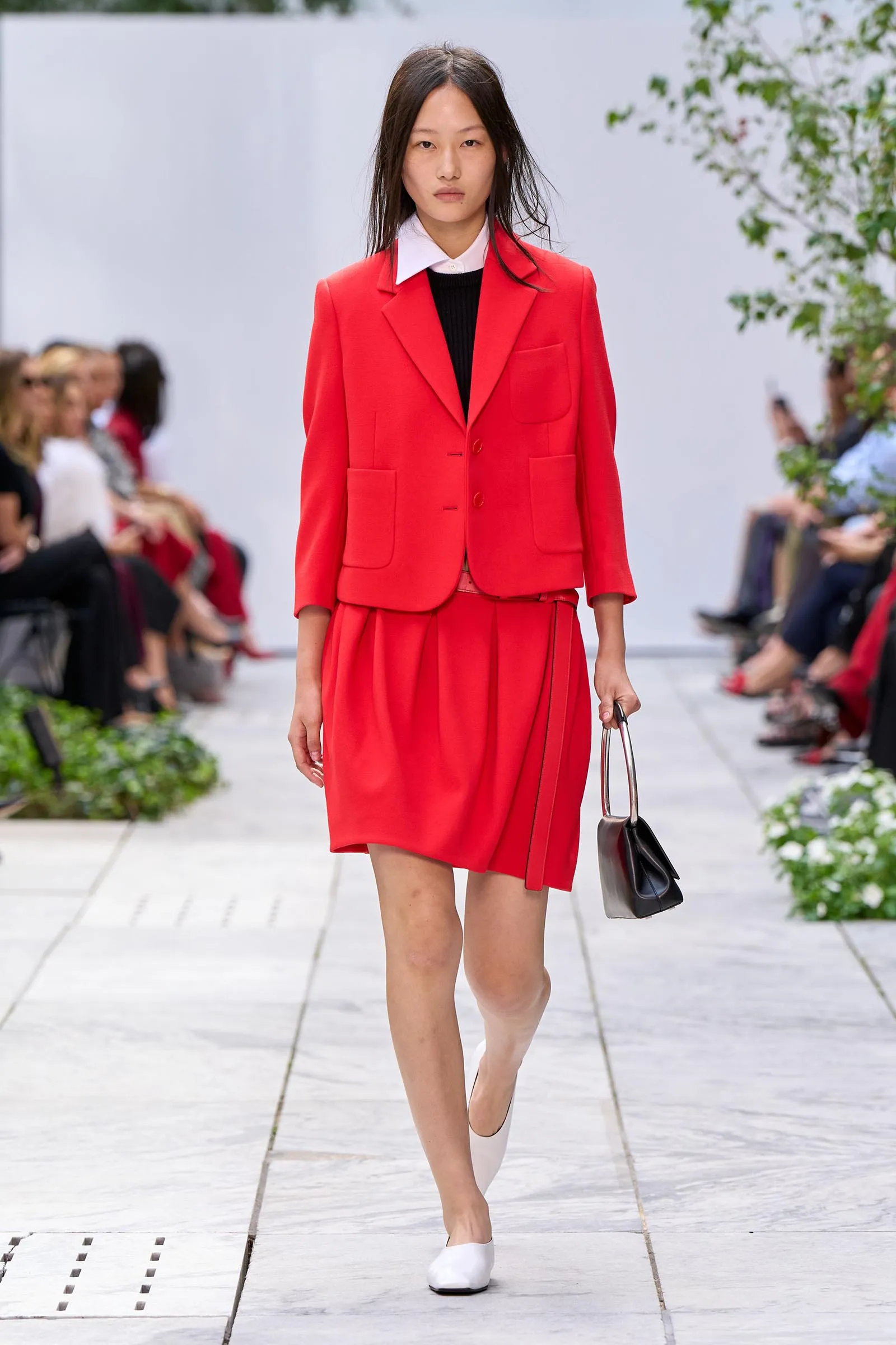 Model walking a runway in a bright red blazer and matching pleated skirt, white shoes, and a small handbag in hand.