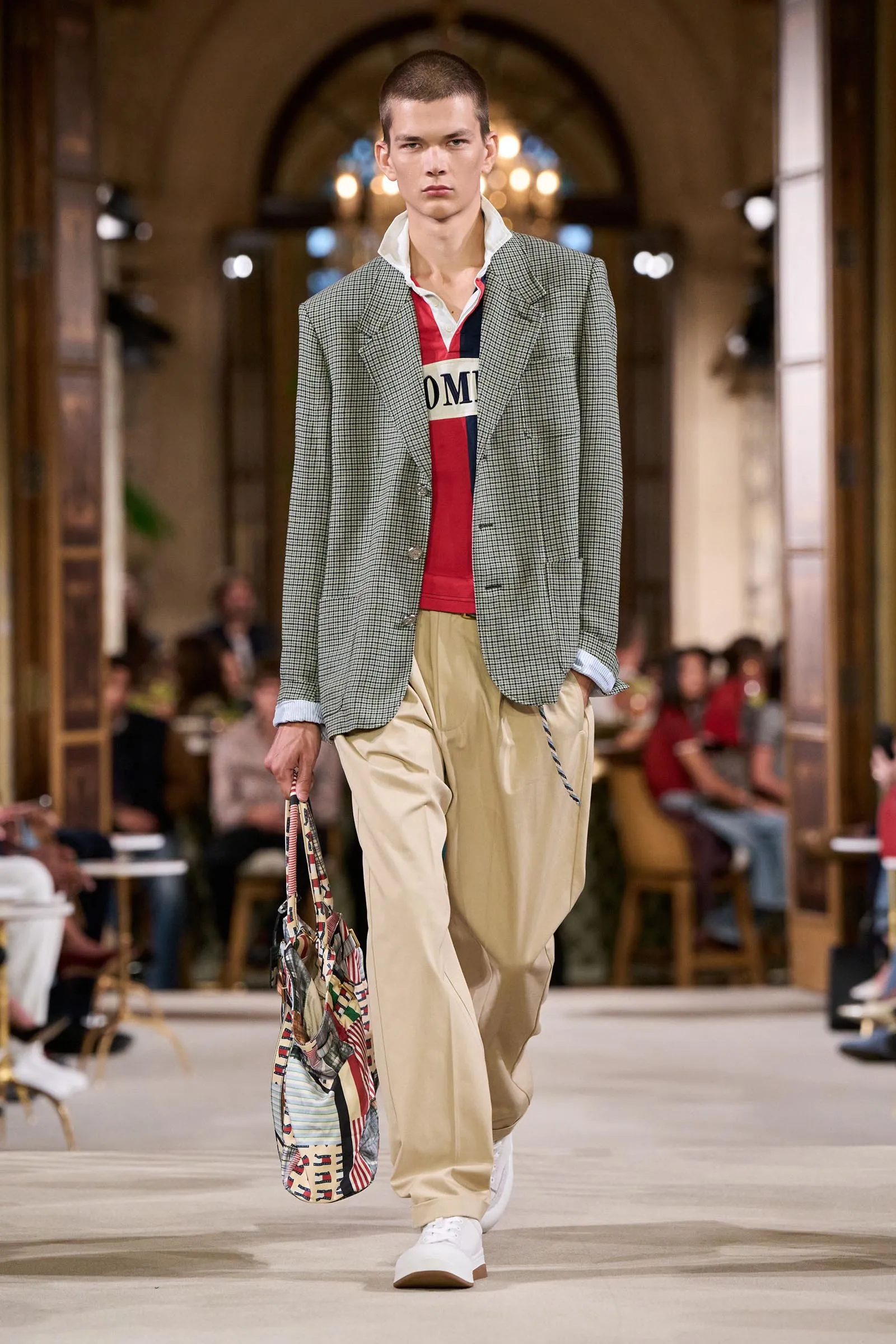 Male model striding down a runway in a gray plaid blazer, red polo, and beige trousers, carrying a patterned tote bag during a fashion show.