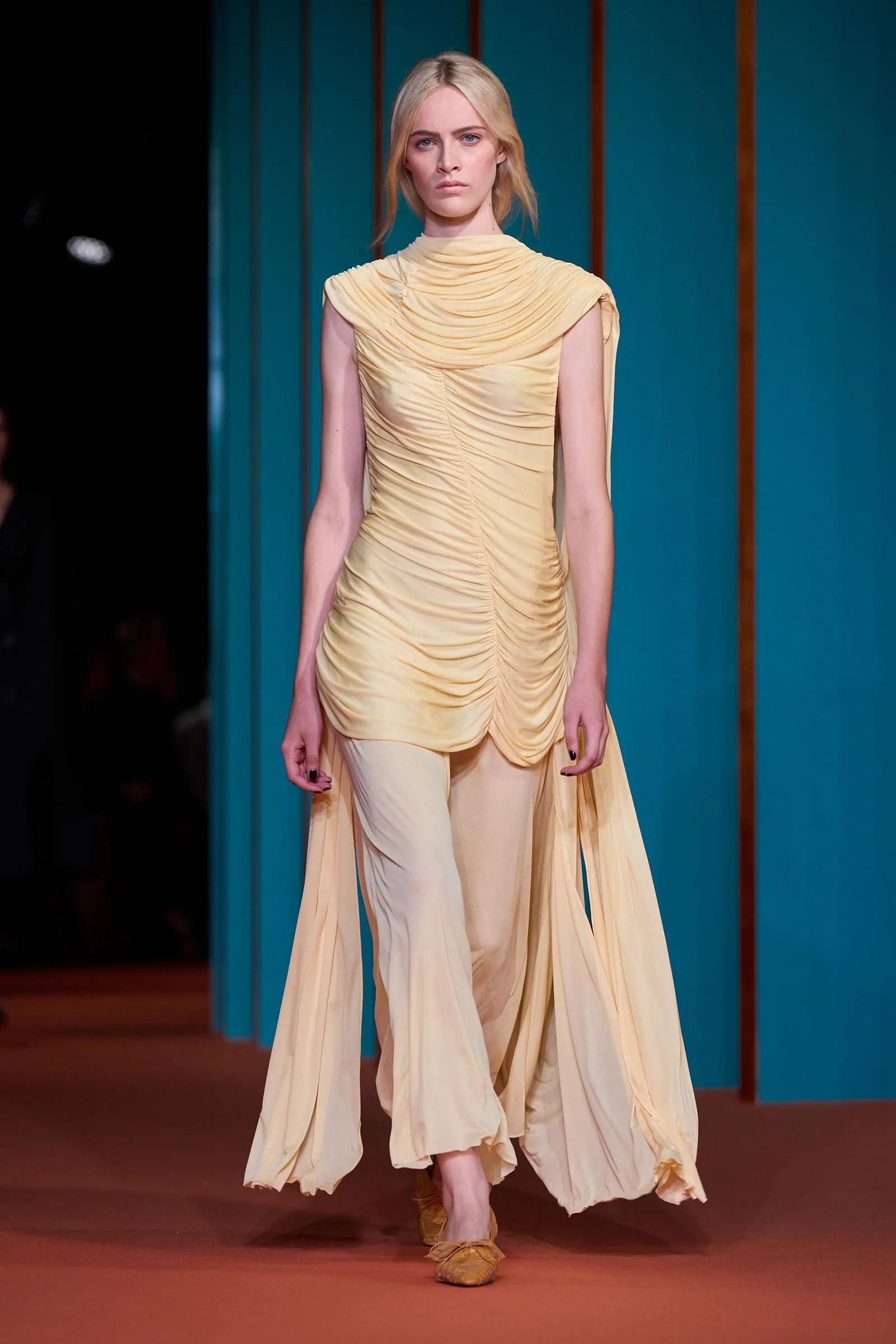 Model walking the runway in a pale yellow, draped gown with cape-like sleeves against a teal backdrop.