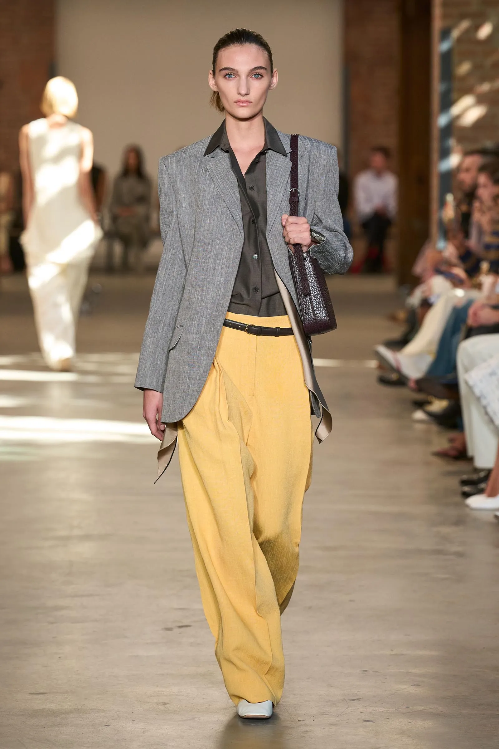 Model on a fashion runway wearing a gray blazer over a dark shirt, yellow wide-leg pants, and carrying a dark brown textured bag across the shoulder.