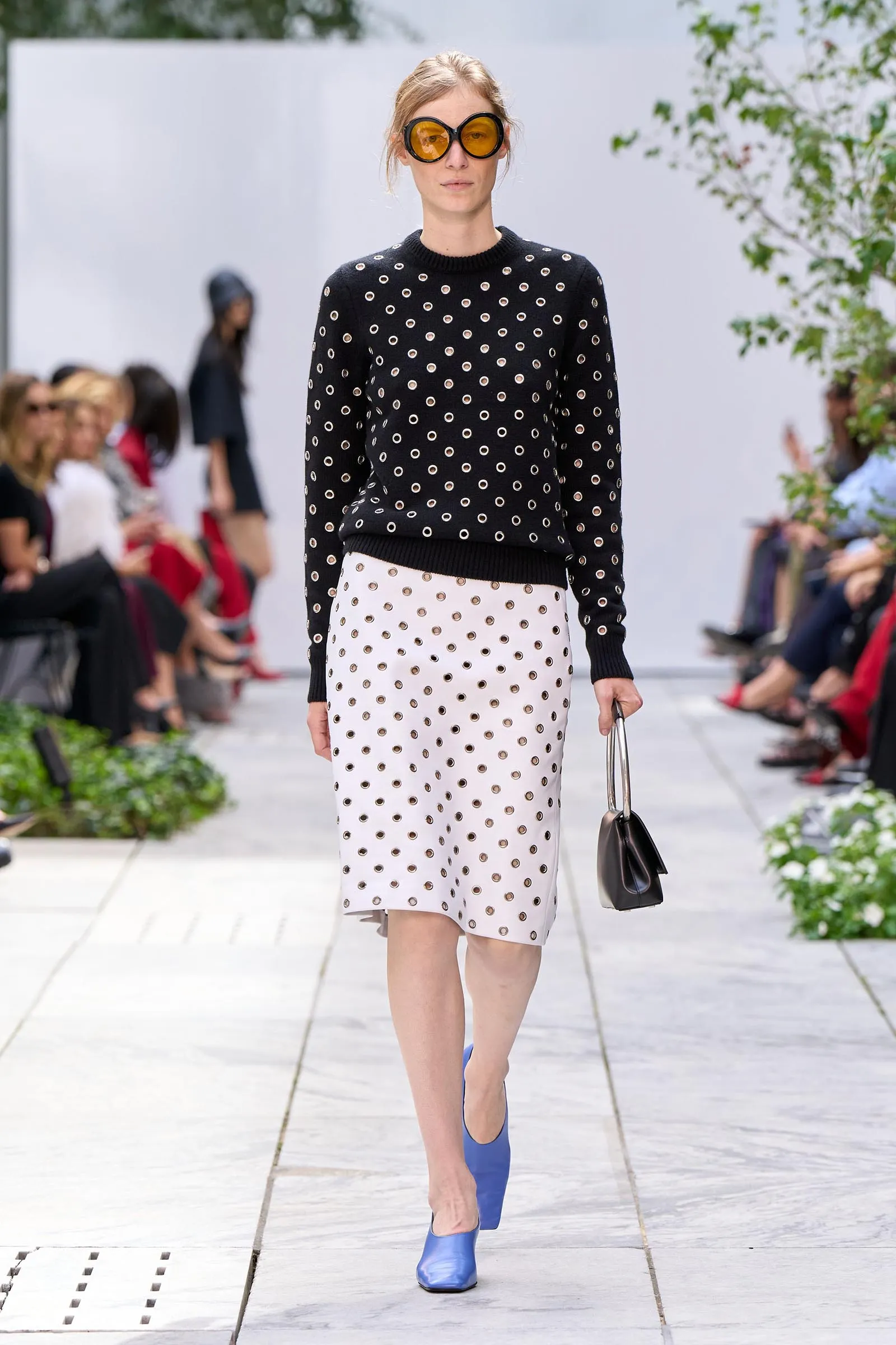 Model walking a fashion show outdoors in a black perforated sweater with circular cutouts, a white dotted midi skirt, blue shoes, orange sunglasses, and a small black bag.