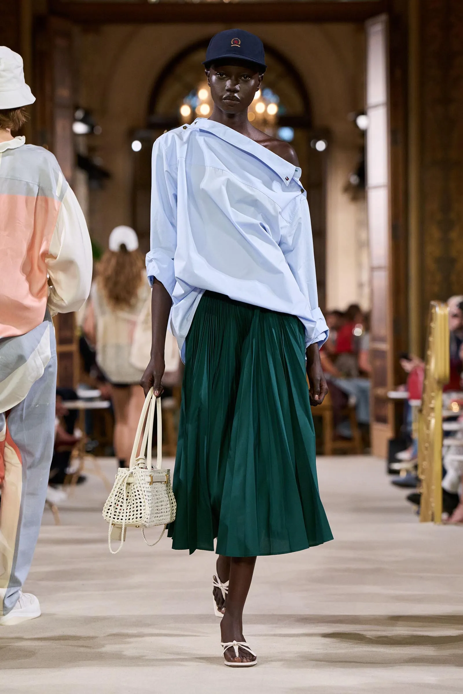 Model walks the runway in a light blue oversized shirt, green pleated skirt, navy cap, white sandals, and a white woven bag.
