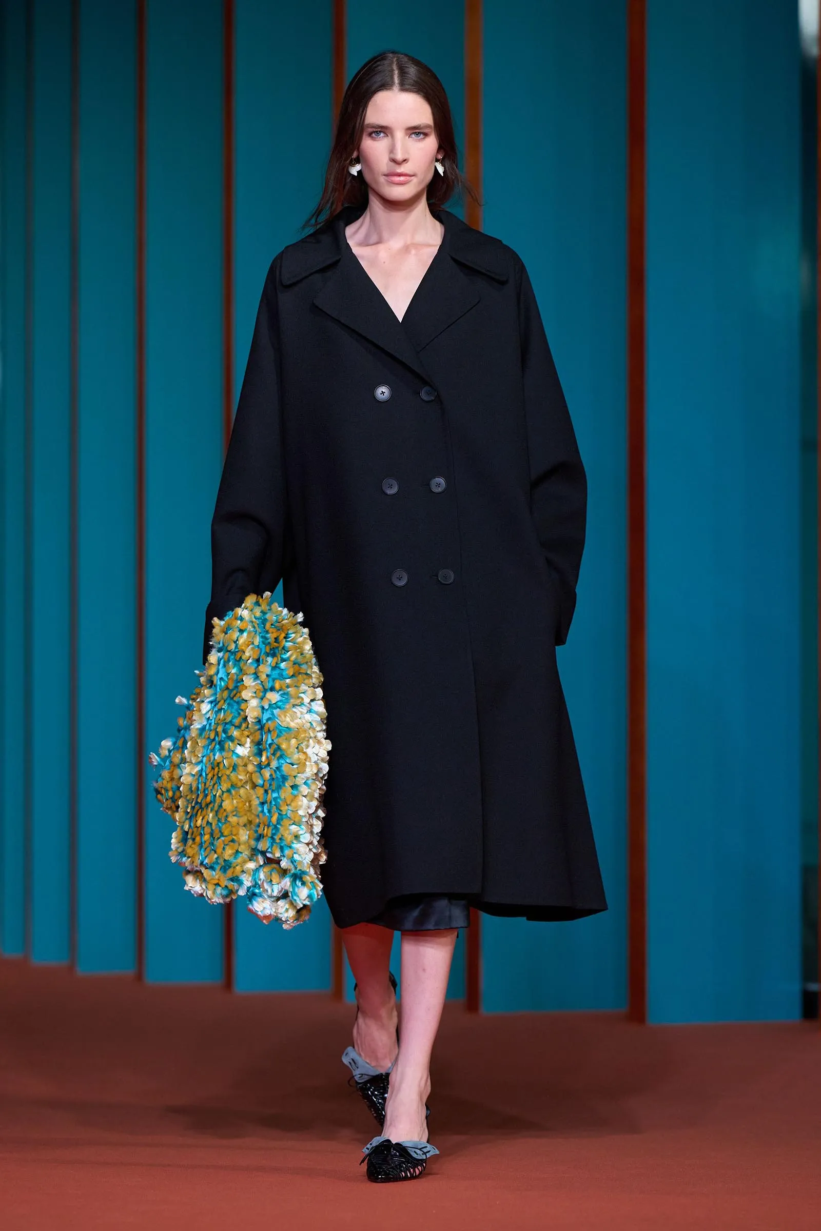 Model in a black double‑breasted coat walking on a red carpet, holding a blue‑yellow floral bouquet, against teal paneled backdrop.