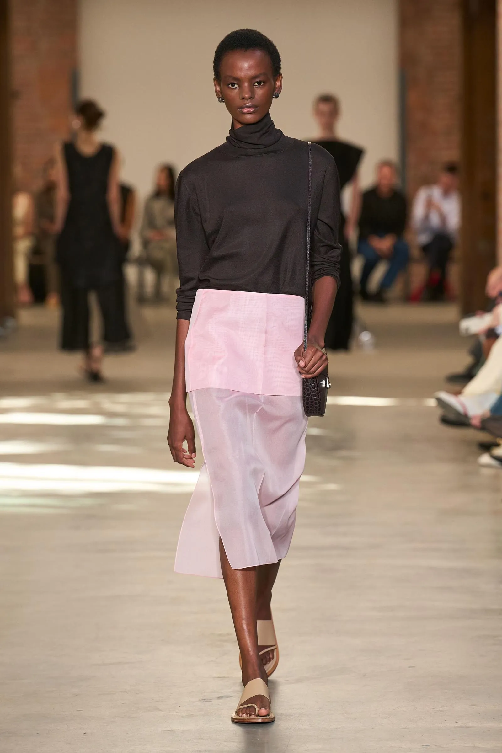 Model walking the runway in a black turtleneck top and pale pink skirt, carrying a small black handbag, beige sandals.