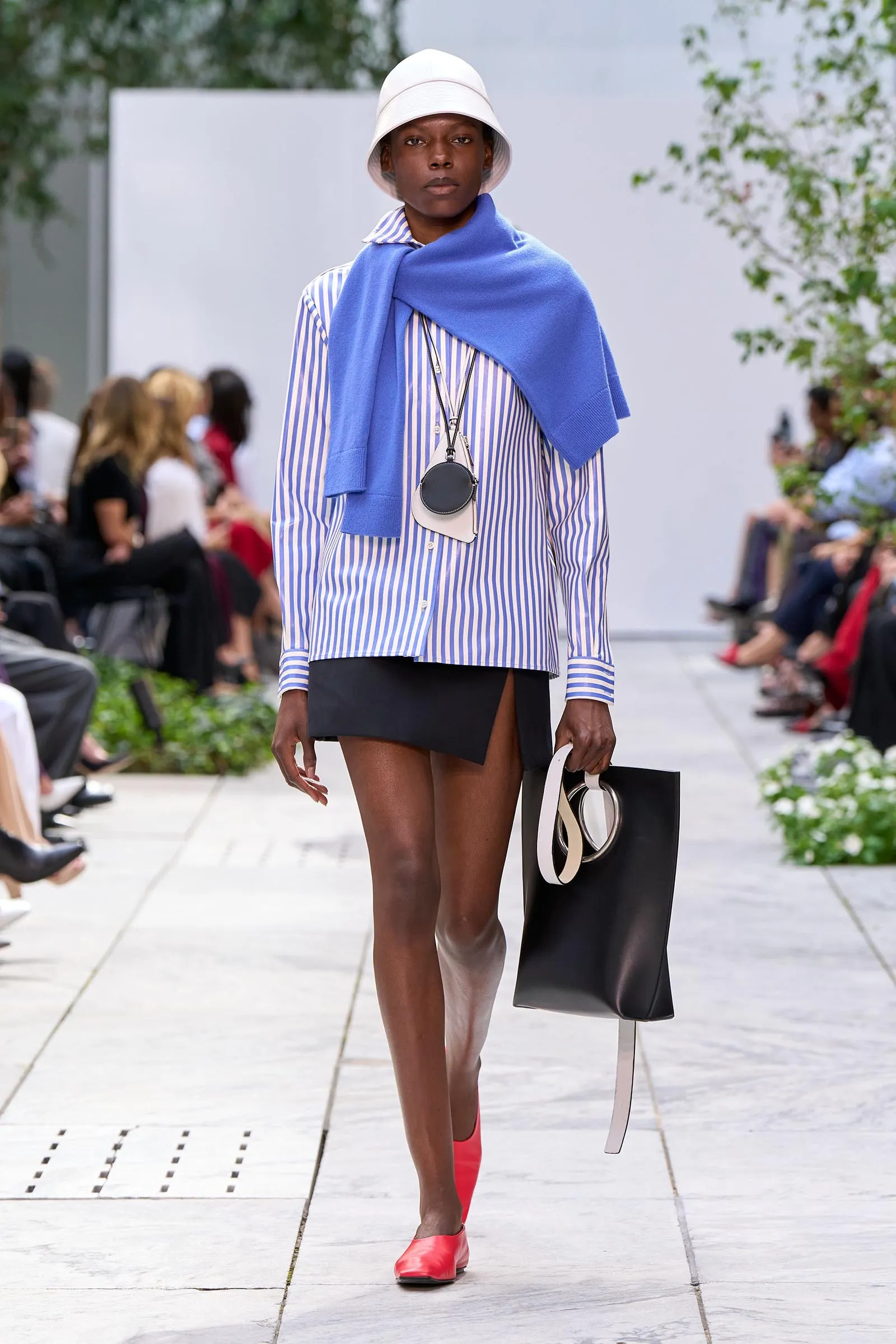 Model on a runway wearing a blue and white striped shirt with a bright blue scarf draped over shoulders, white bucket hat, black mini skirt, red shoes, and a large black tote bag with beige handles.