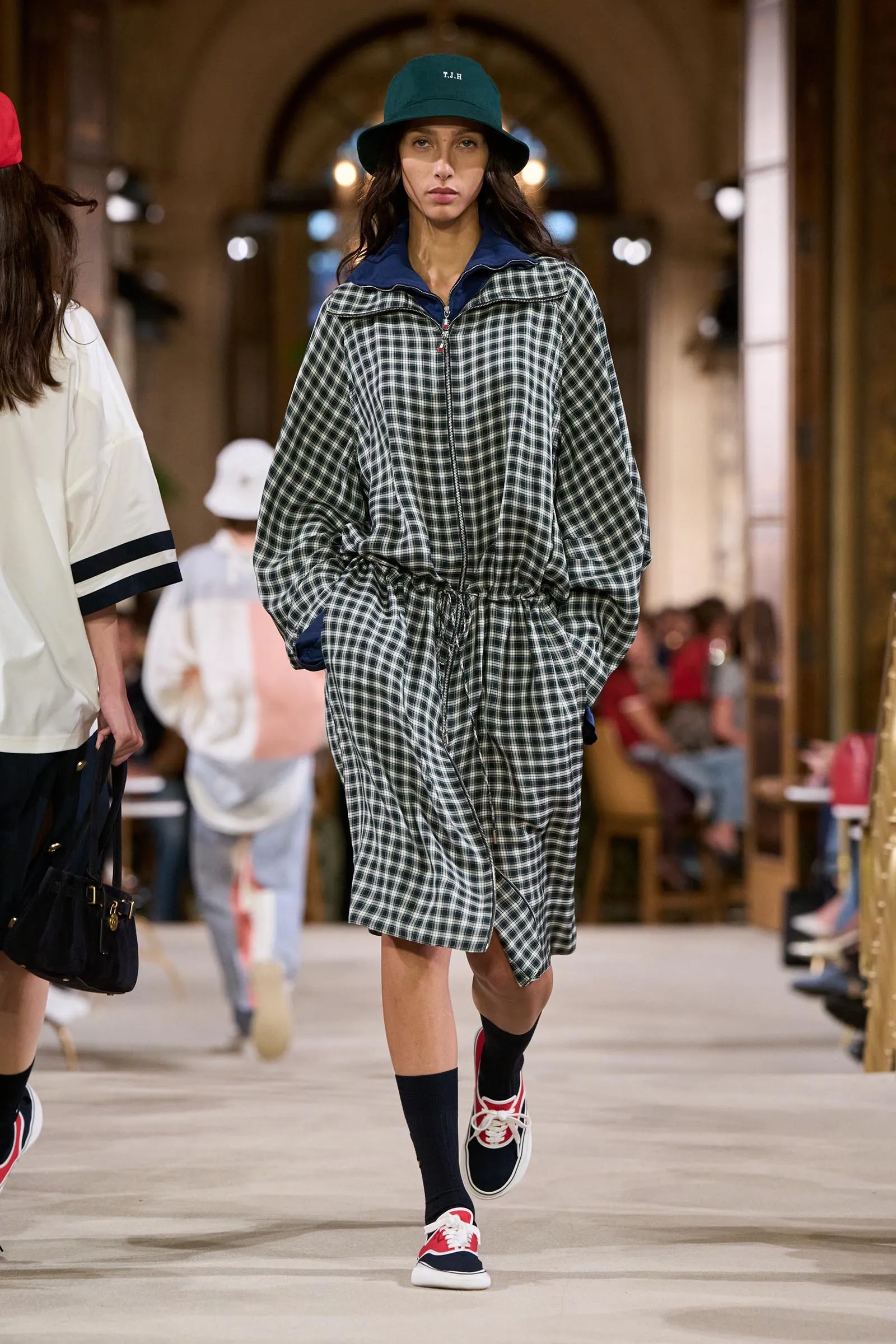 Female model walks a fashion show runway in a black-and-white plaid oversized jacket, blue collar, green bucket hat, and red sneakers. Audience blurred in background.