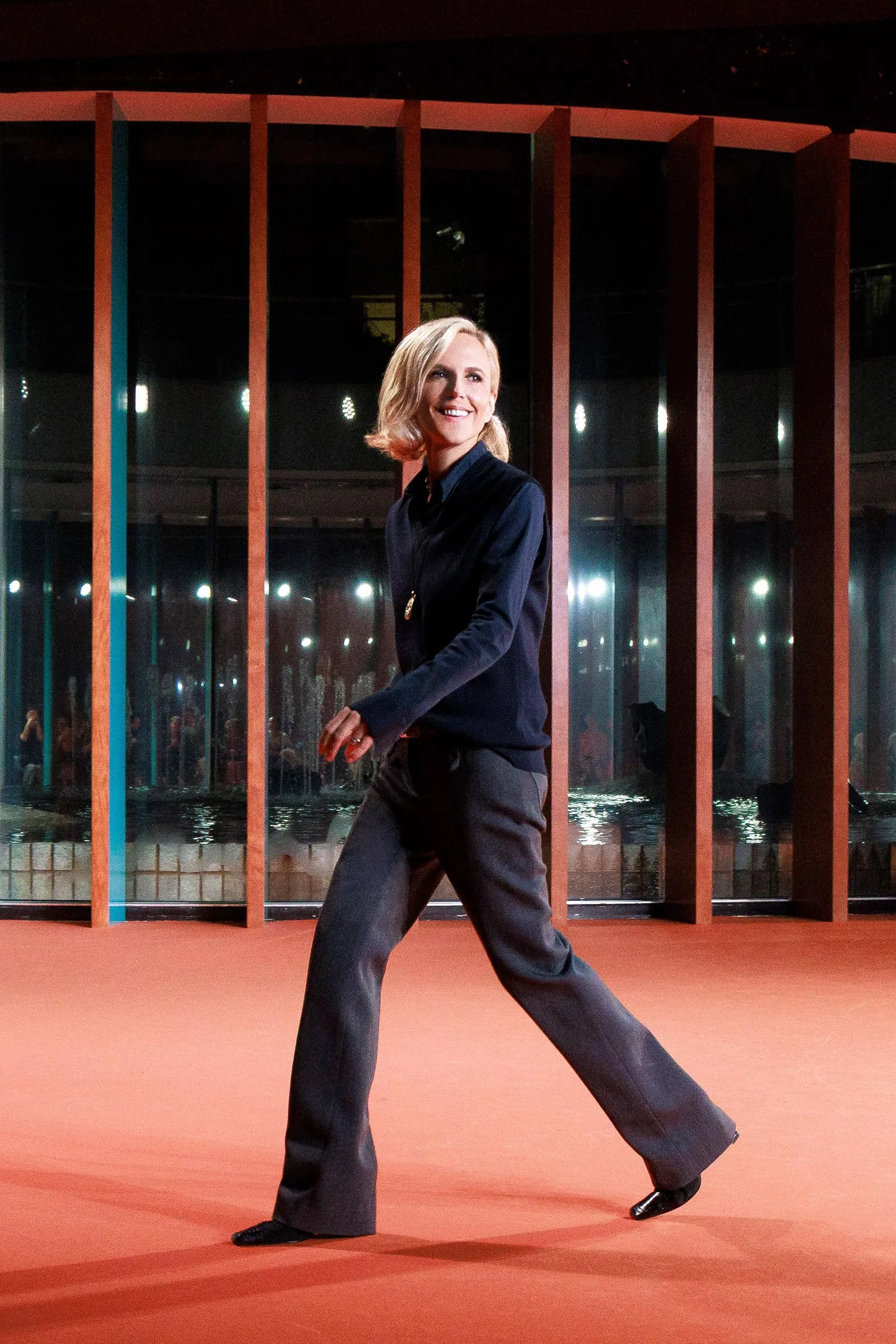 Woman in a dark blue outfit walks across an orange floor, smiling, with vertical wooden beams behind her.