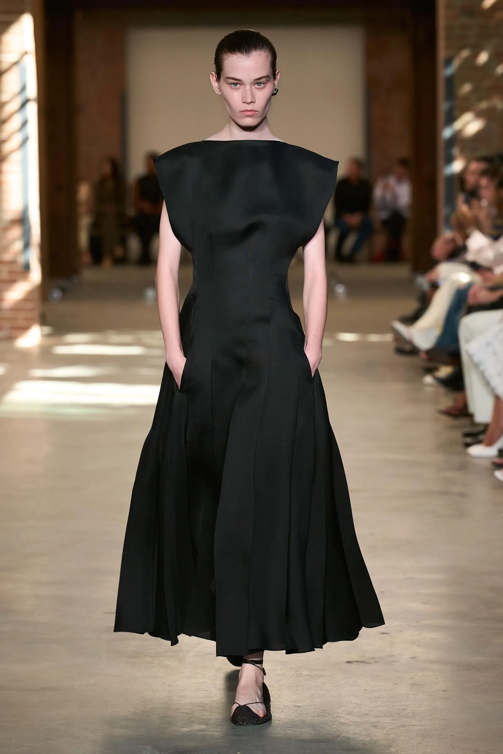 Model on runway wearing a long black sculptural gown with broad shoulders and a fitted waist.