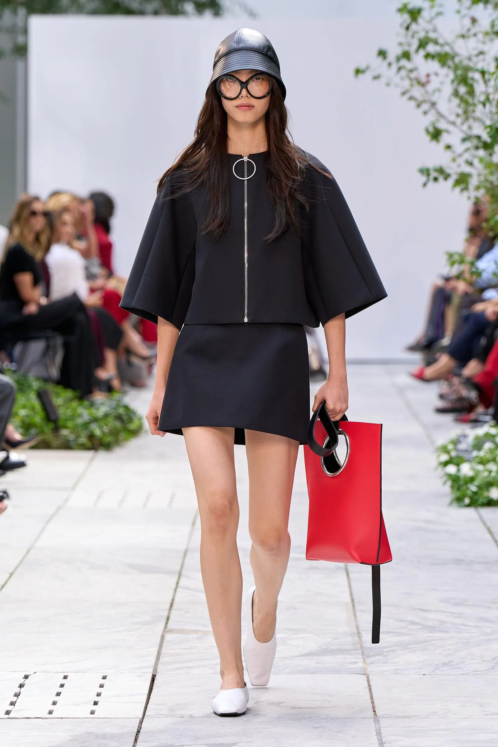 Model walking a runway in a black cape-like outfit with a silver zipper and circular ring pull, pairing oversized glasses and a metallic helmet, carrying a red tote bag.