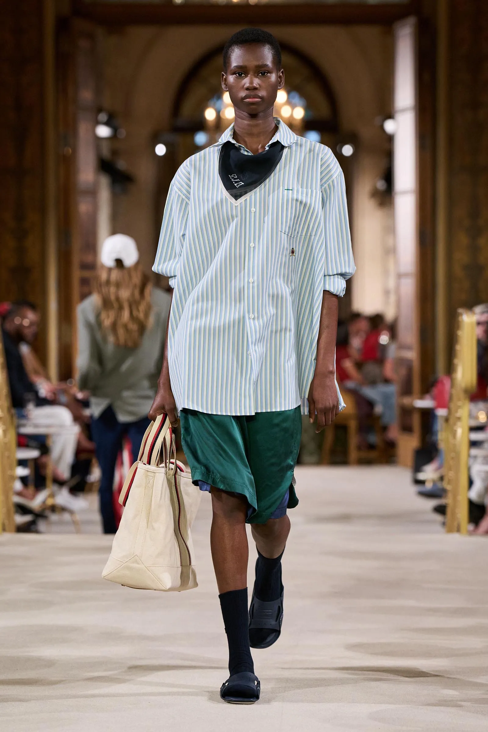 Model walking a fashion runway in a light blue and white striped oversized shirt, green shorts, black scarf, and dark slides, carrying a beige tote bag.