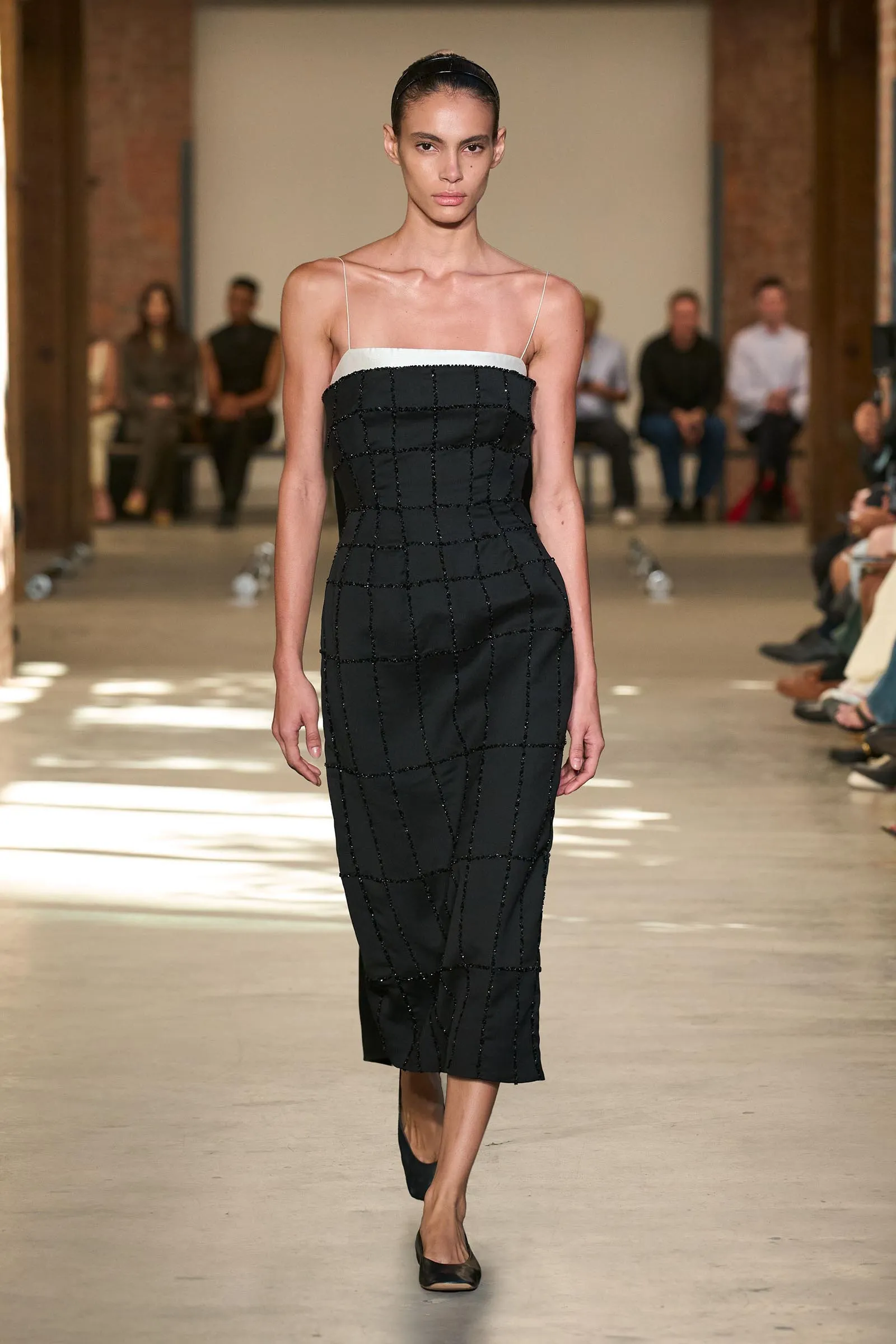 Runway model walking in a black grid-patterned dress with a white neckline and thin spaghetti straps.