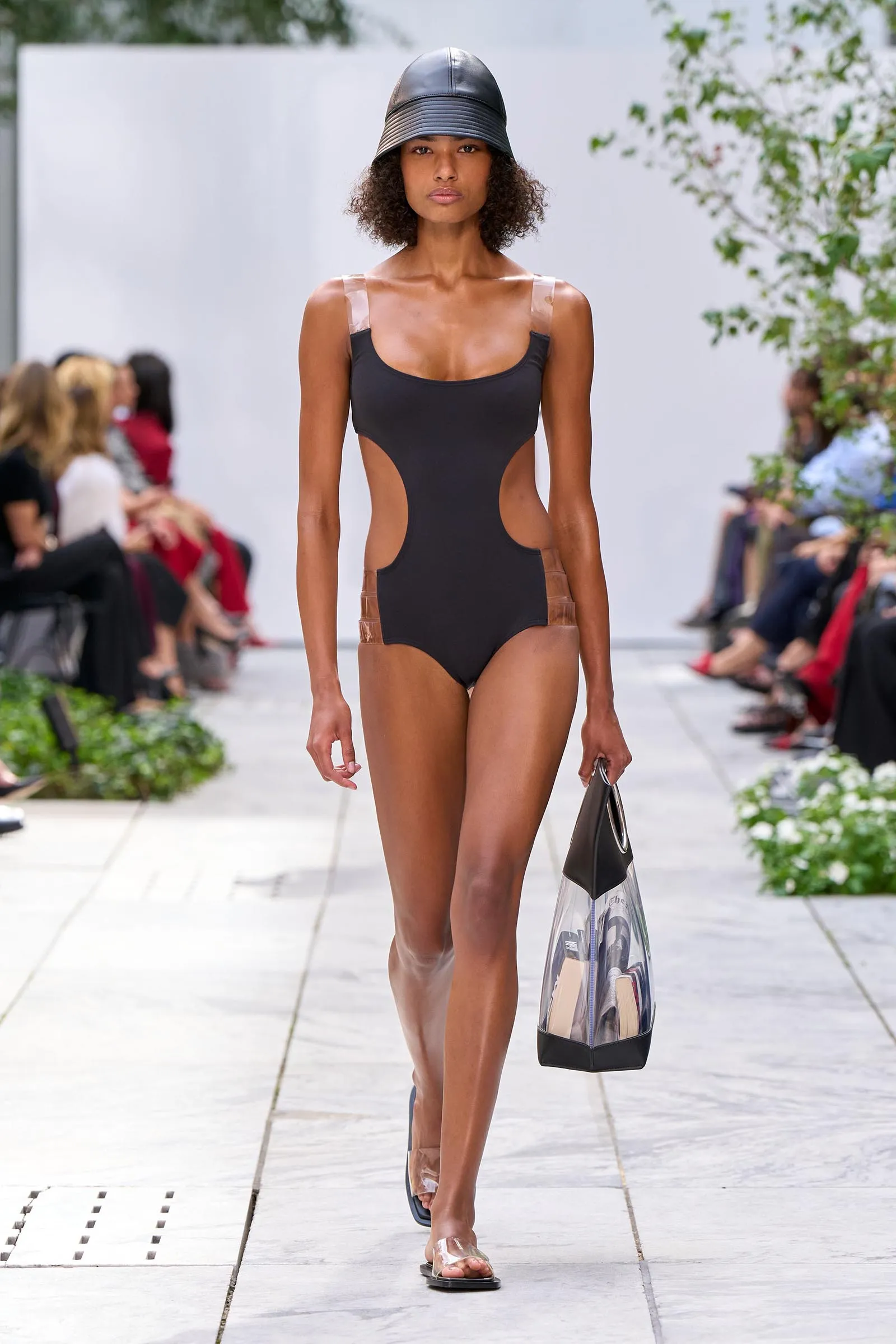 Model walks a fashion runway in a black cut-out one-piece swimsuit and metallic bucket hat, carrying a clear tote bag with items inside.