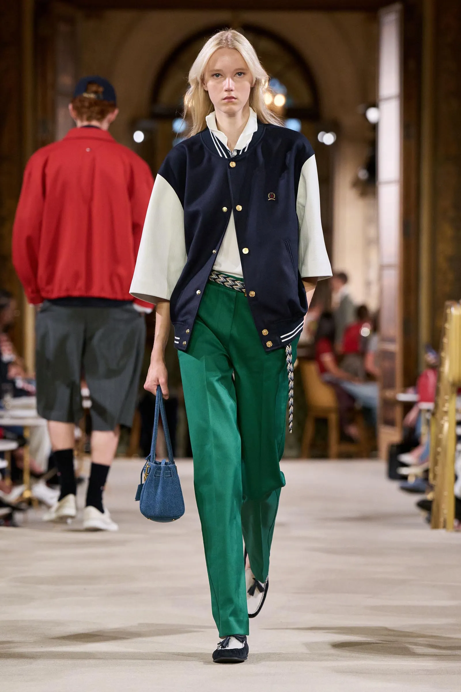 Model walking a fashion runway in a navy varsity jacket, white sleeves, green trousers, and a blue handbag, indoors.