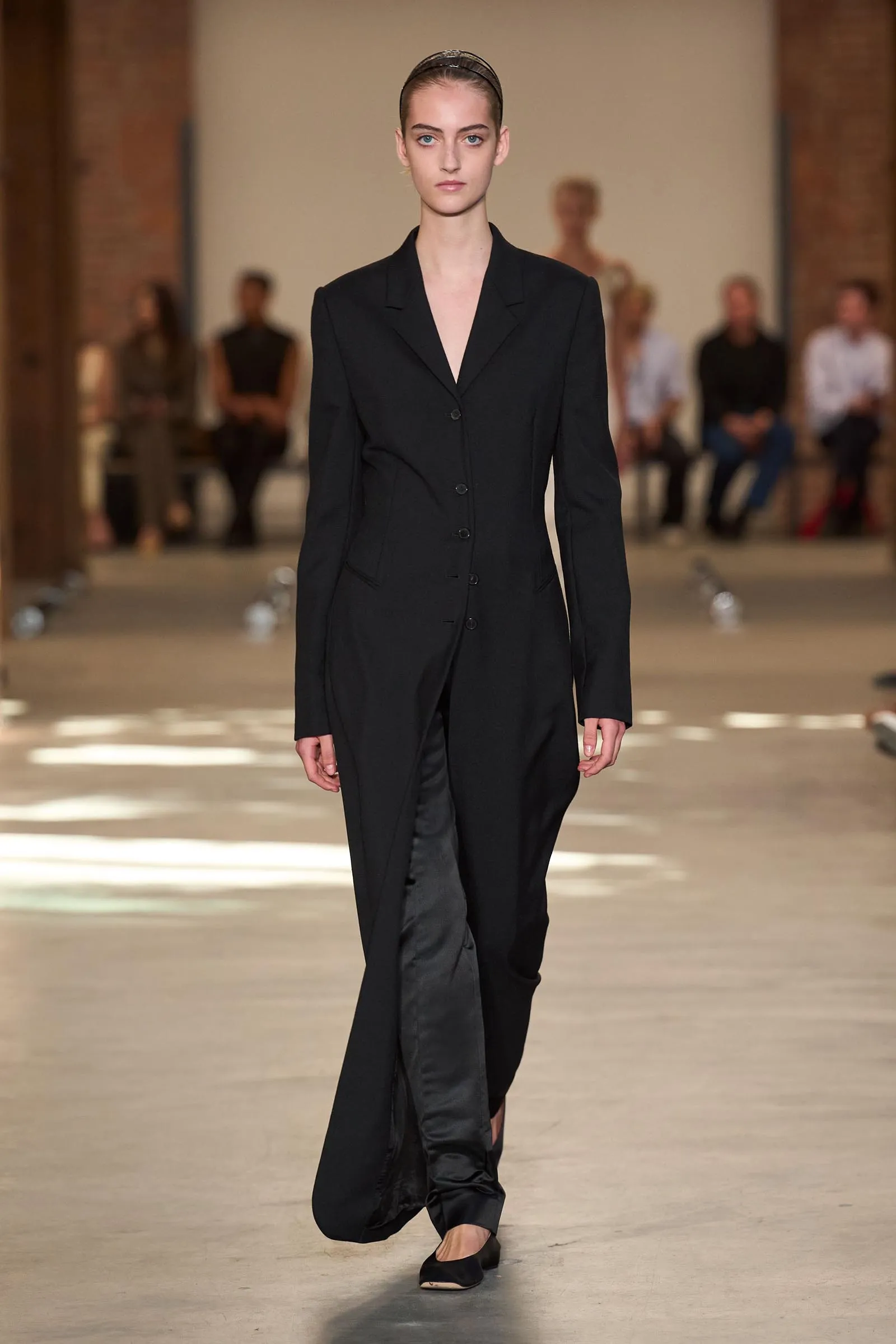Model walking the runway in a black tailored suit with a long blazer and matching pants; headband in hair, neutral expression, blurred audience in the background.