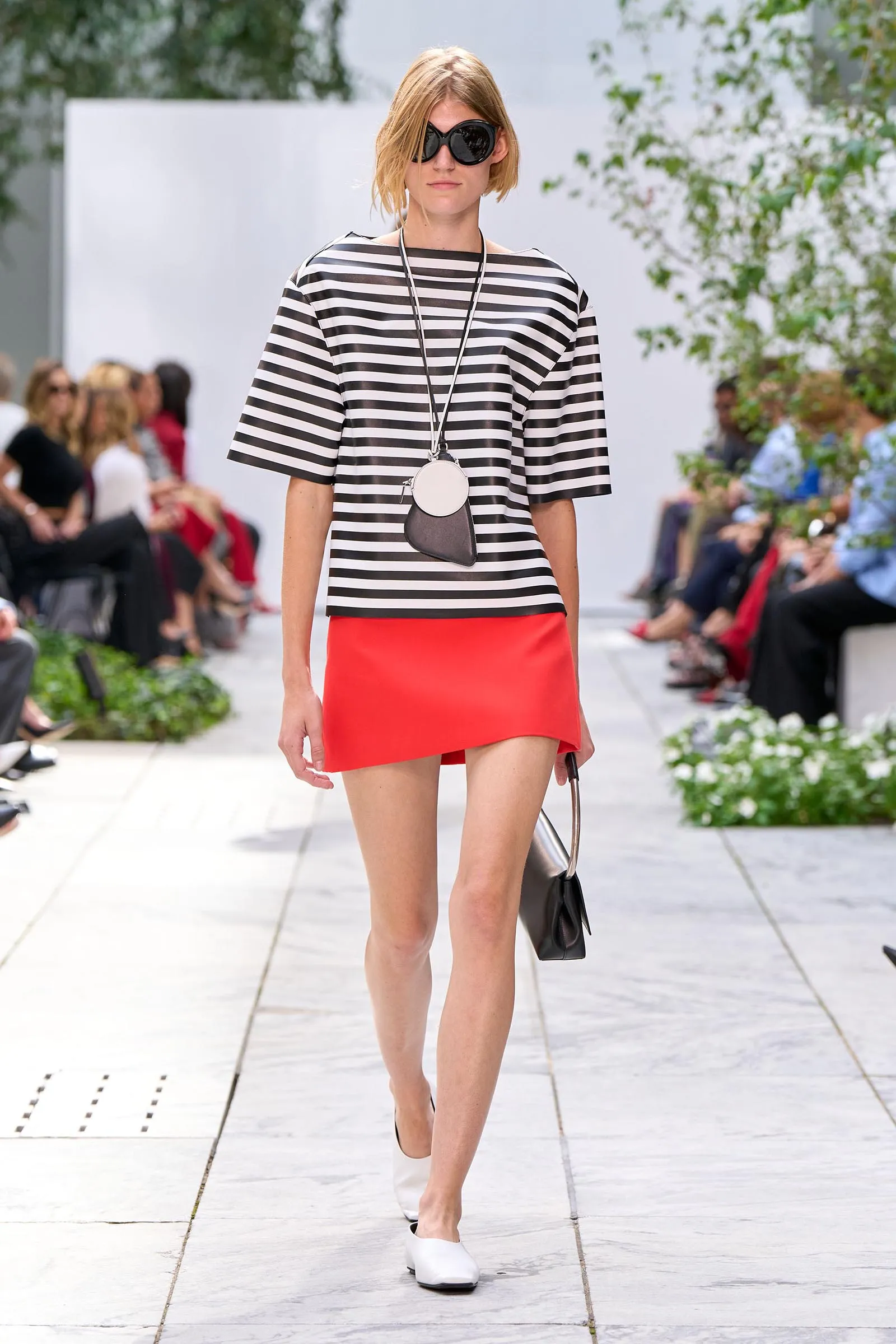 Model walking a runway in a black-and-white striped top, red mini skirt, white flats, oversized sunglasses, and a circular white pendant bag on a long strap.