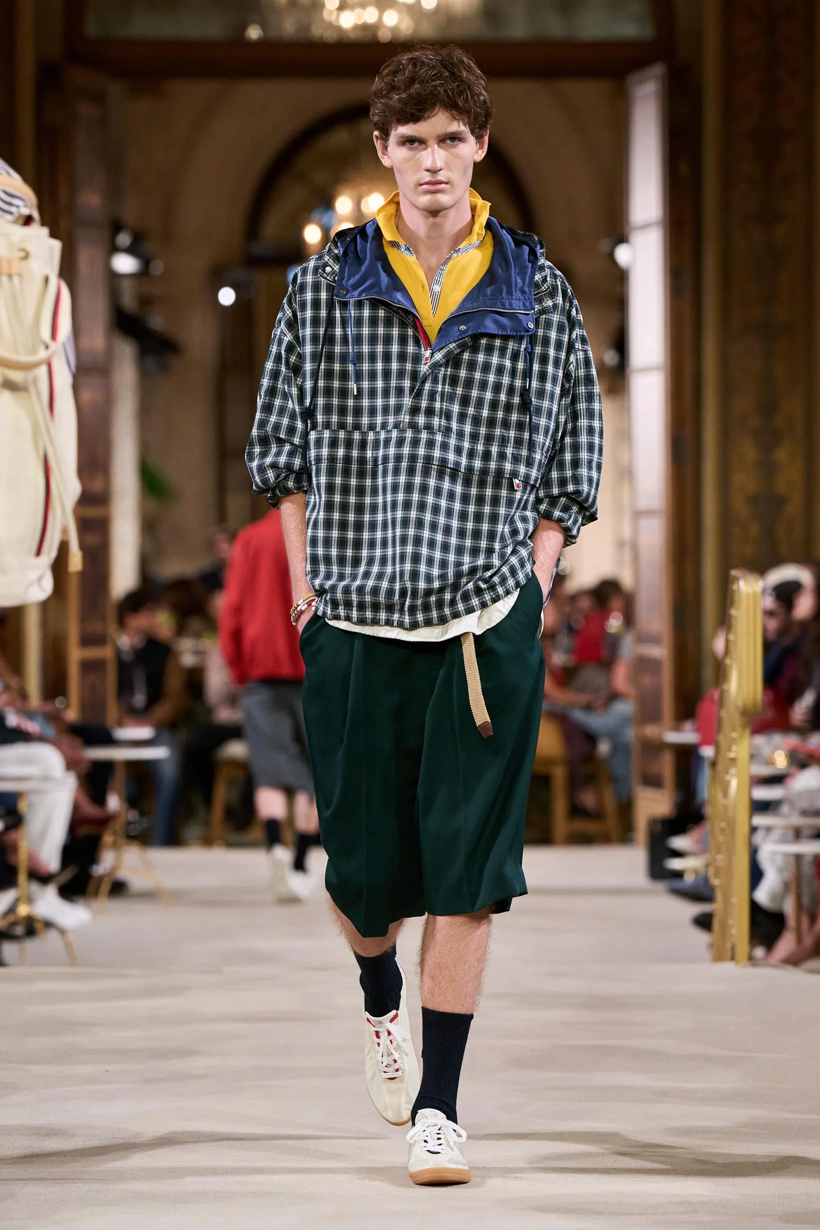 Male model walks a runway wearing a green plaid windbreaker over a yellow-collar shirt and dark green shorts with white sneakers on a fashion show.