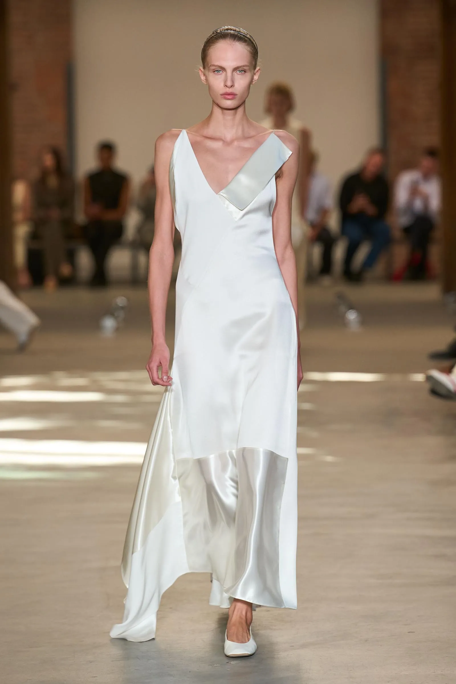 Model walking a fashion show in a white satin gown with a plunging V-neck and layered hem, headband in her hair, audience blurred in the background.