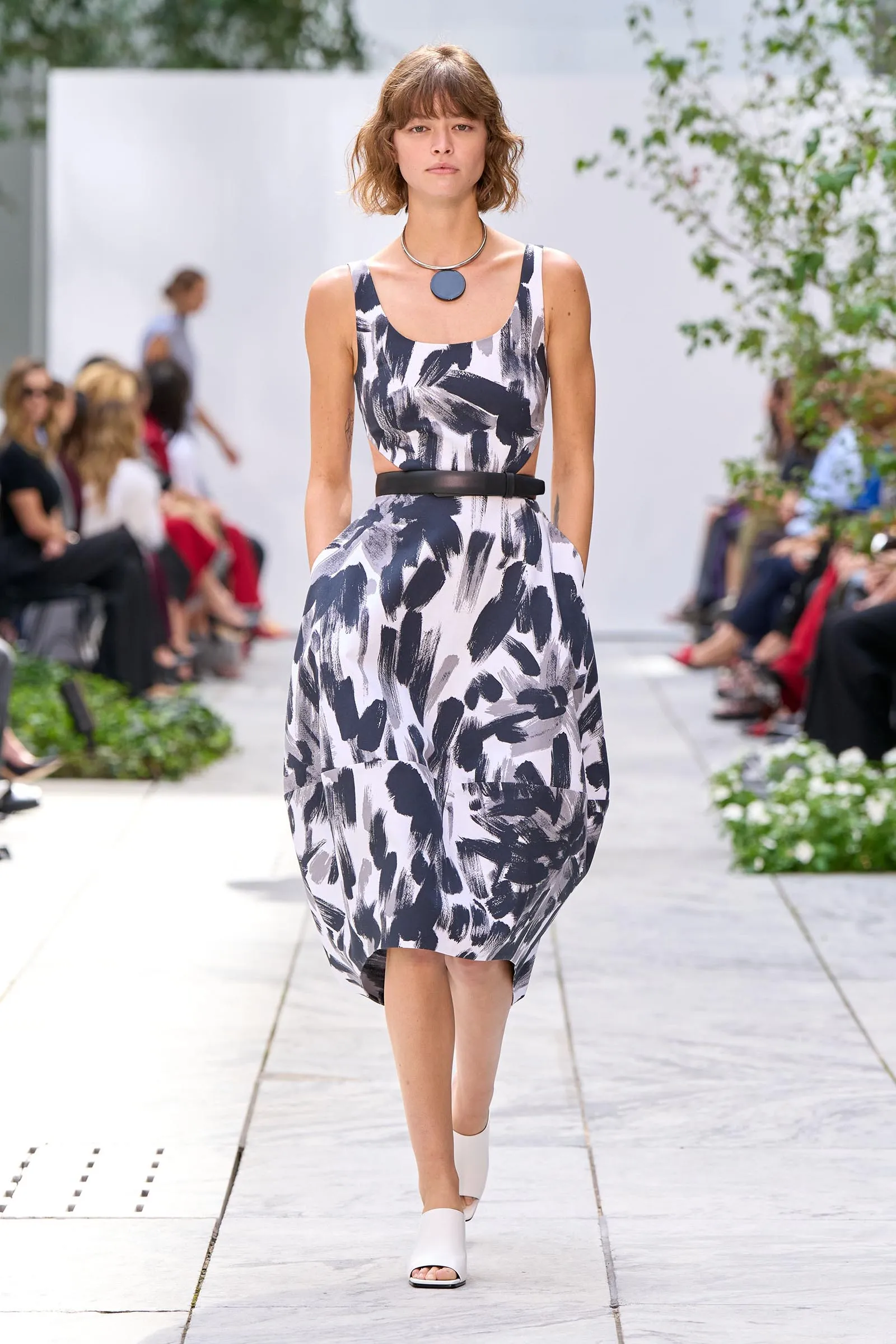 Model walks a fashion runway in a sleeveless black-and-white abstract print dress with a slim black belt and white sandals.
