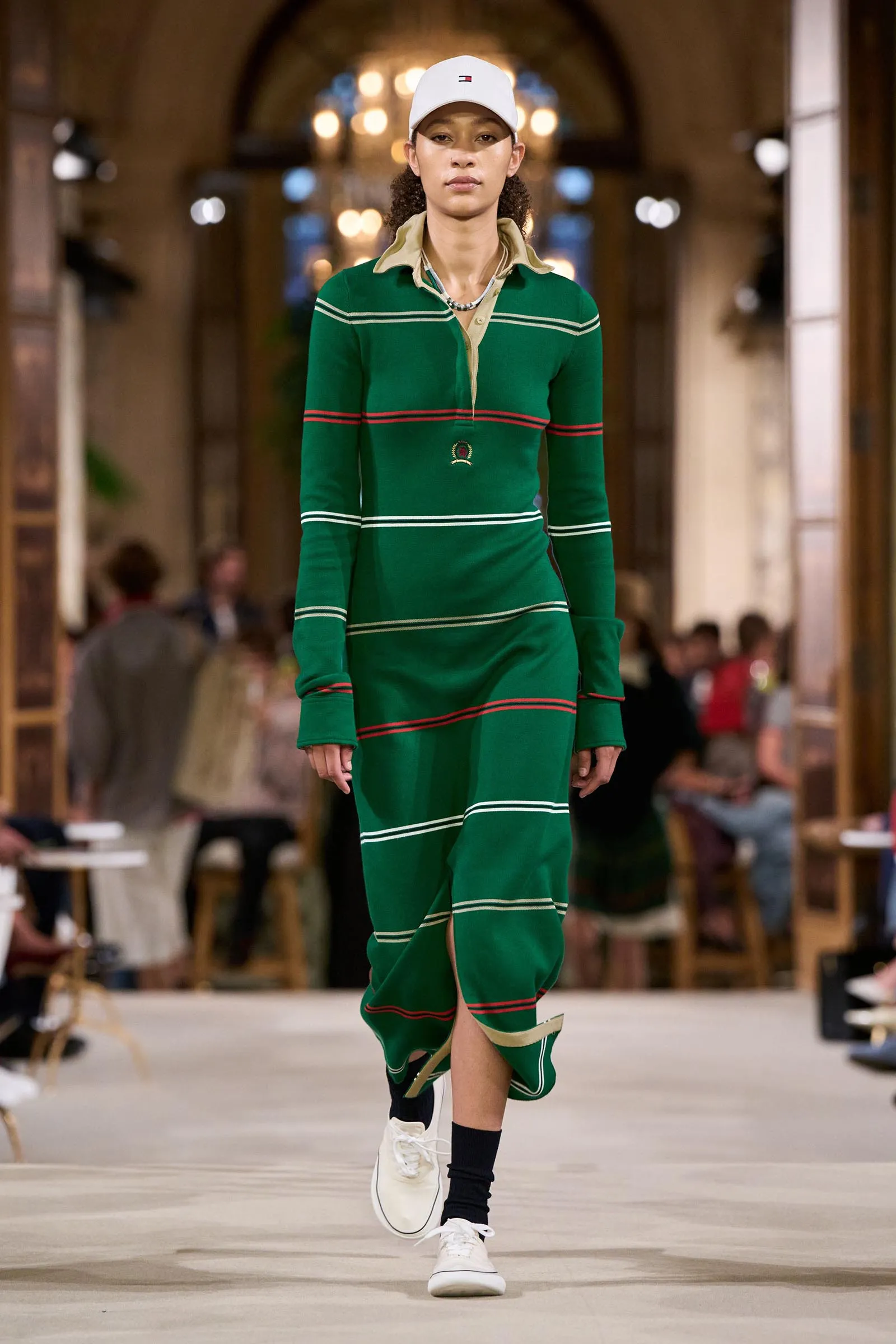 Model walking a green striped dress down a runway, wearing a white cap and sneakers in an indoor fashion show setting (audience blurred).