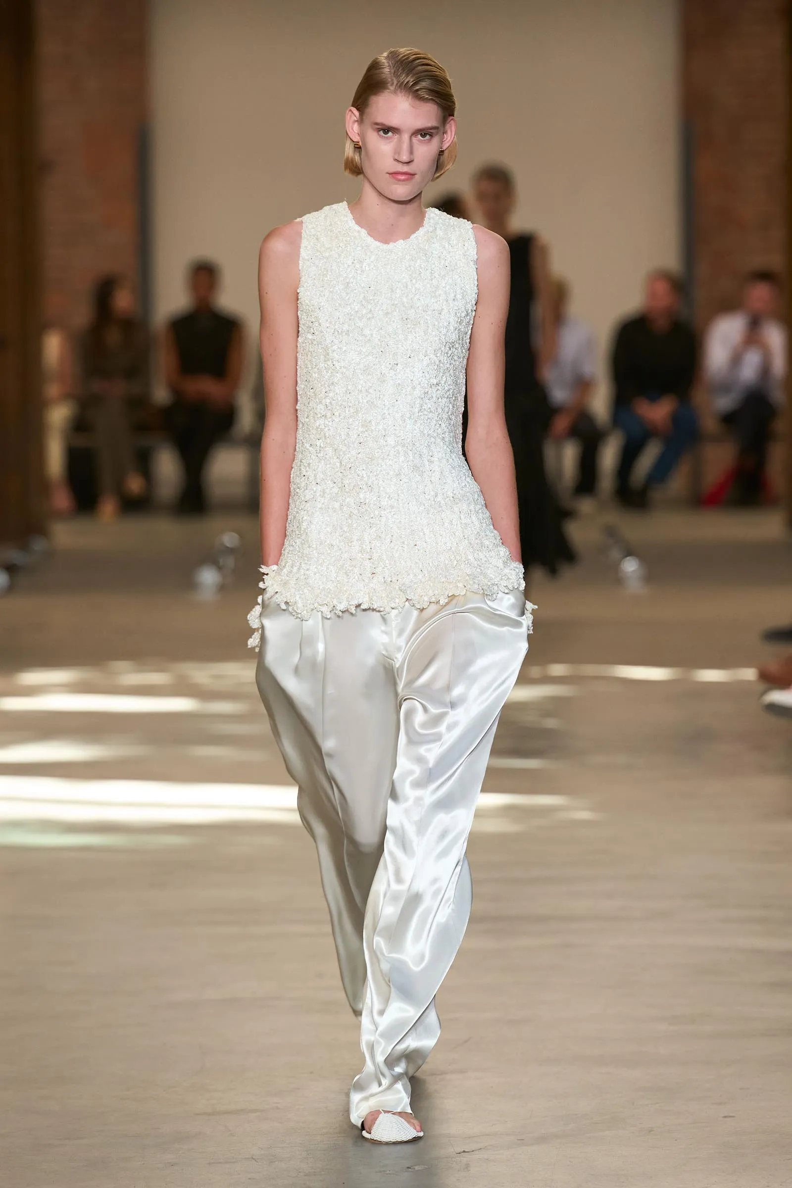 Model walks a runway in a white textured sleeveless top paired with satin pants; audience out of focus in the background.