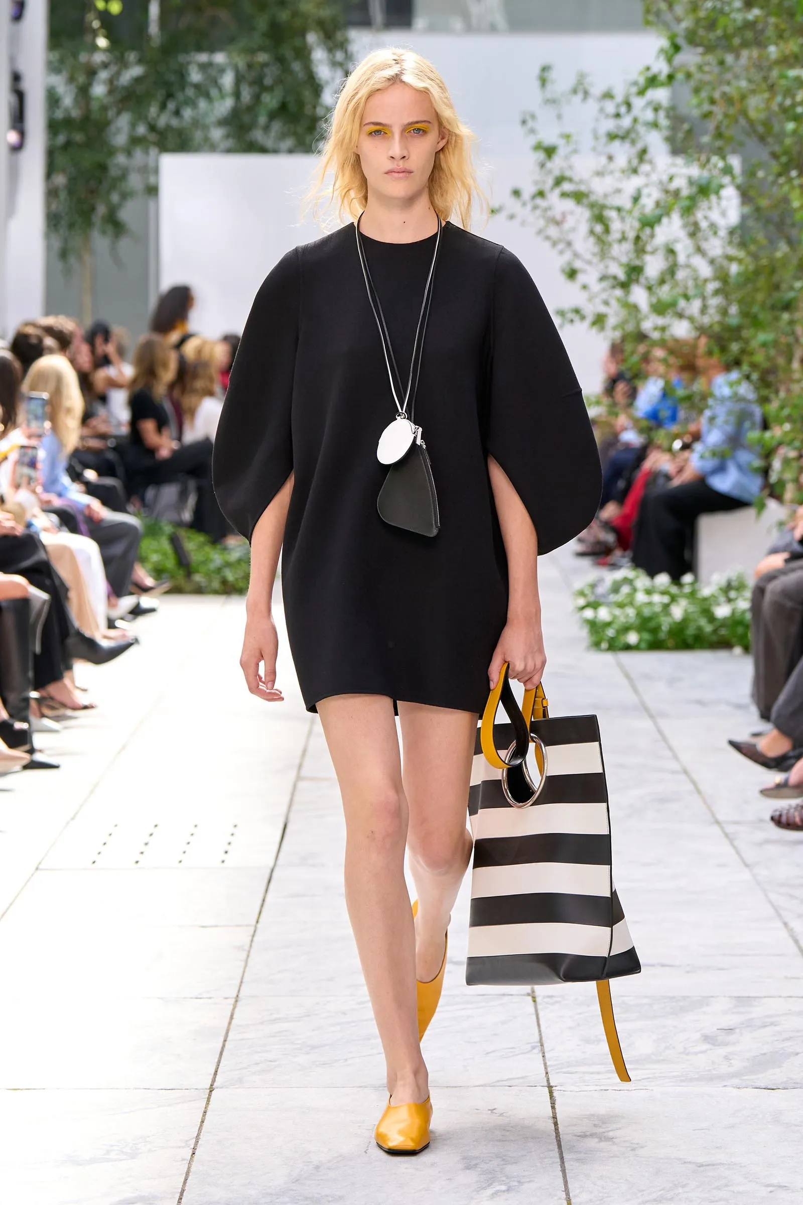 Model walks the runway in a black loose dress, yellow shoes, and a large striped tote bag with a long necklace accessory.