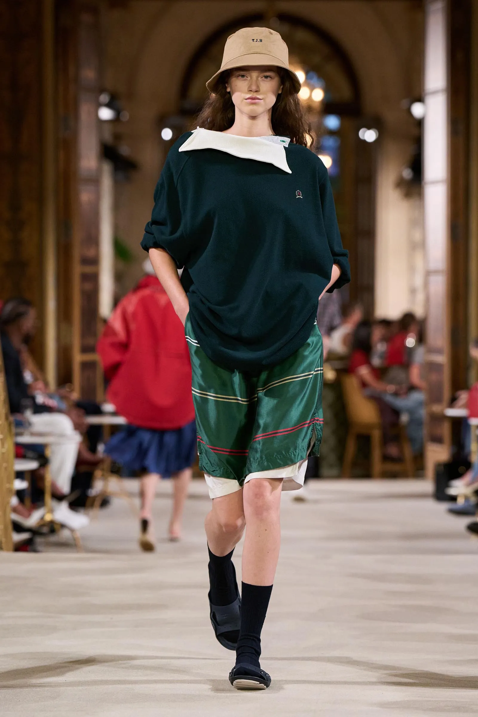 Model on a runway wearing a dark green oversized top with a white collar, green striped shorts, black socks, sandals, and a beige bucket hat.