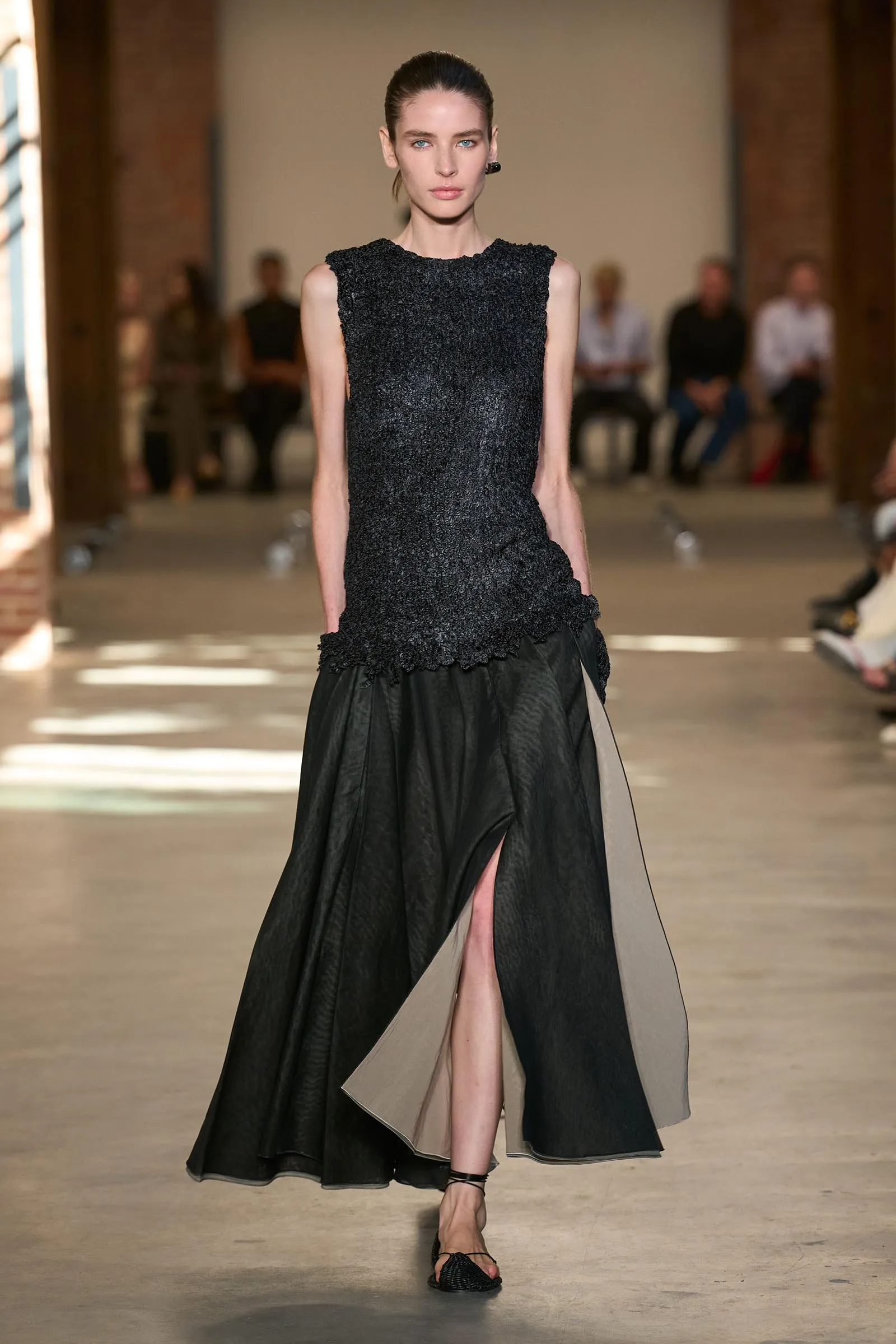 Model walking a runway in a black textured sleeveless top and a two-tone black and beige floor-length skirt with a front slit, audience blurred in the background.