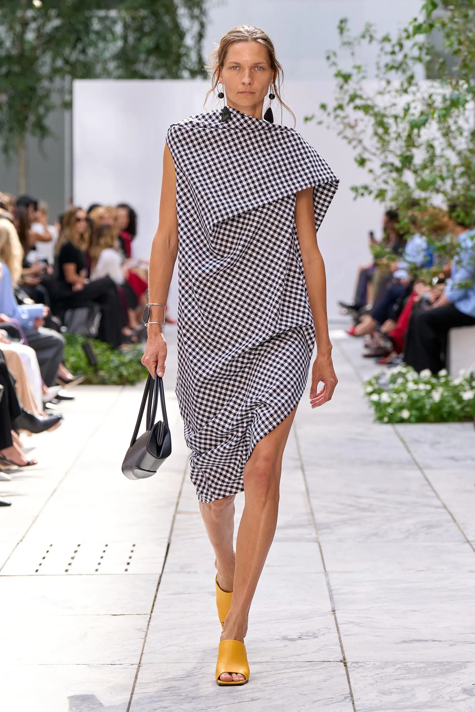 Model wearing a black-and-white gingham draped dress walks a runway outdoors, carrying a black handbag.