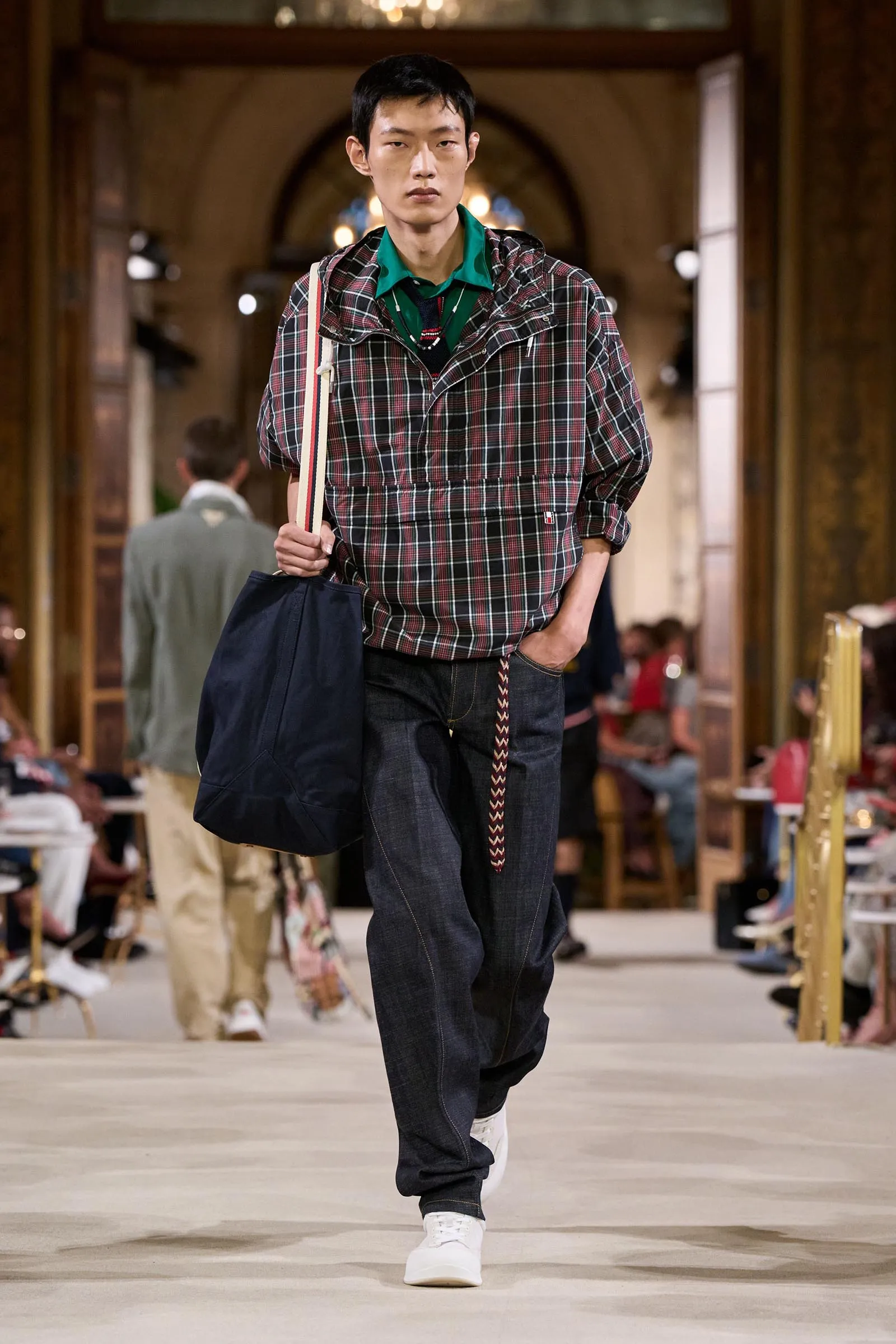 Male model walks the runway in a red plaid jacket over a green shirt, dark jeans, white sneakers, carrying a navy bag on his shoulder.