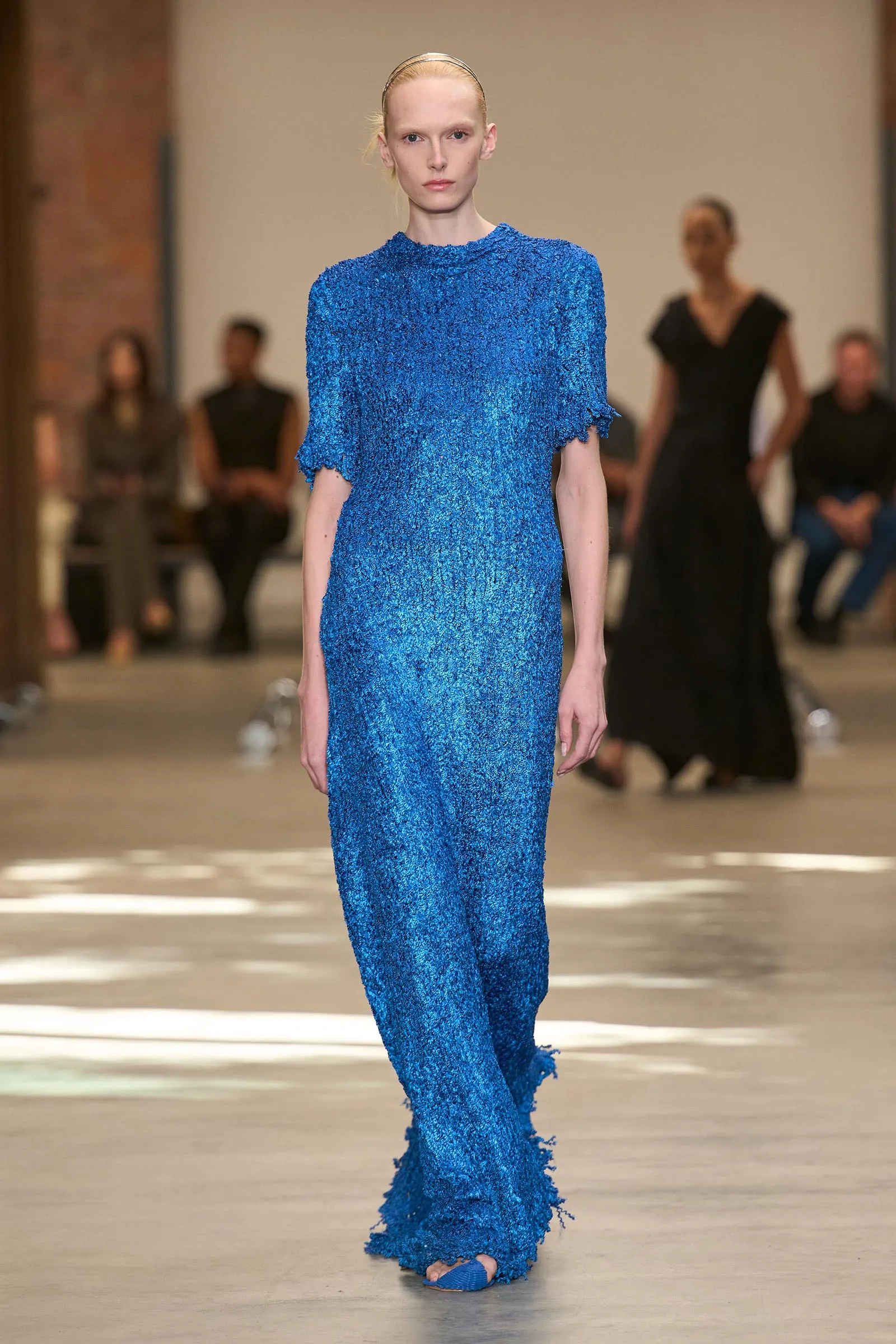 Model walking a cobalt-blue, sequined gown down a runway with blurred spectators in the background.